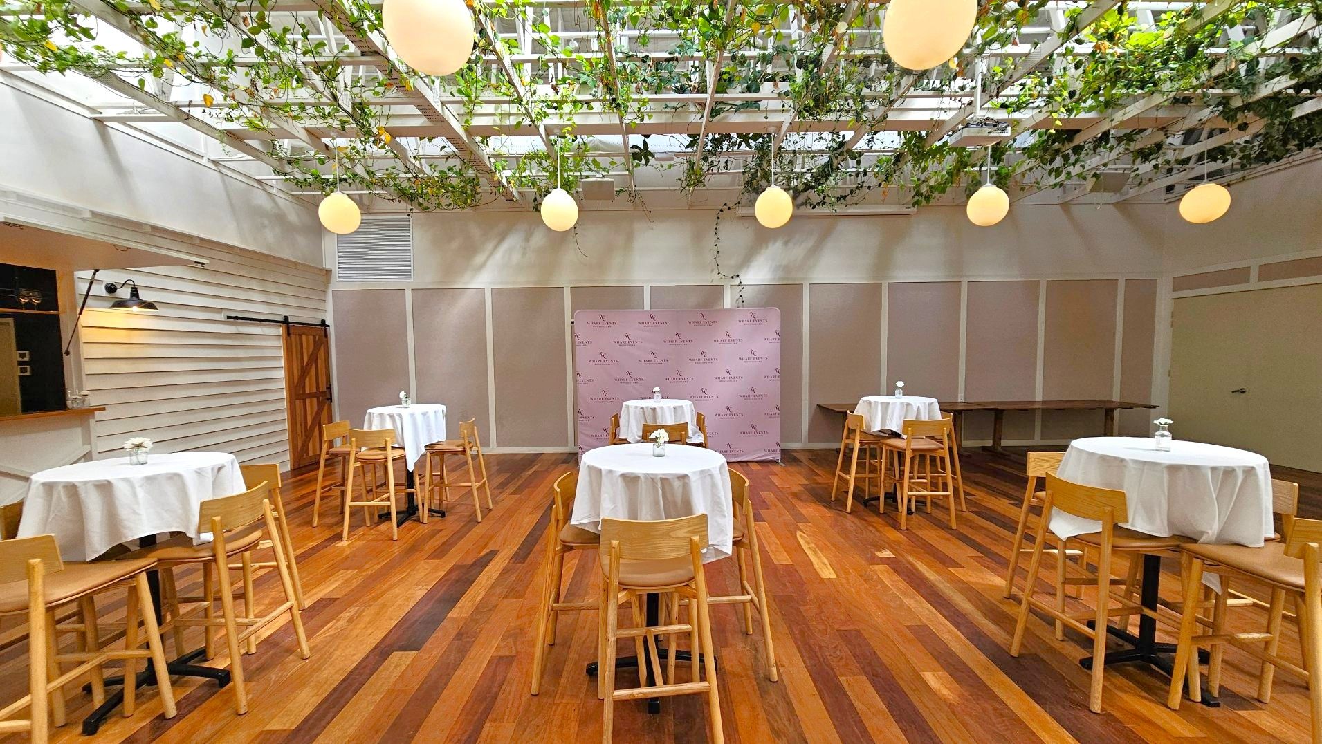 A spacious room with wooden flooring features several round tables covered with white tablecloths, each surrounded by wooden chairs. The ceiling is adorned with hanging plants and spherical pendant lights. A backdrop with a repeating logo pattern stands against one wall. A sliding barn door and a small bar area with a wall-mounted lamp are visible on the left. The walls are light-coloured, creating an open and airy atmosphere.