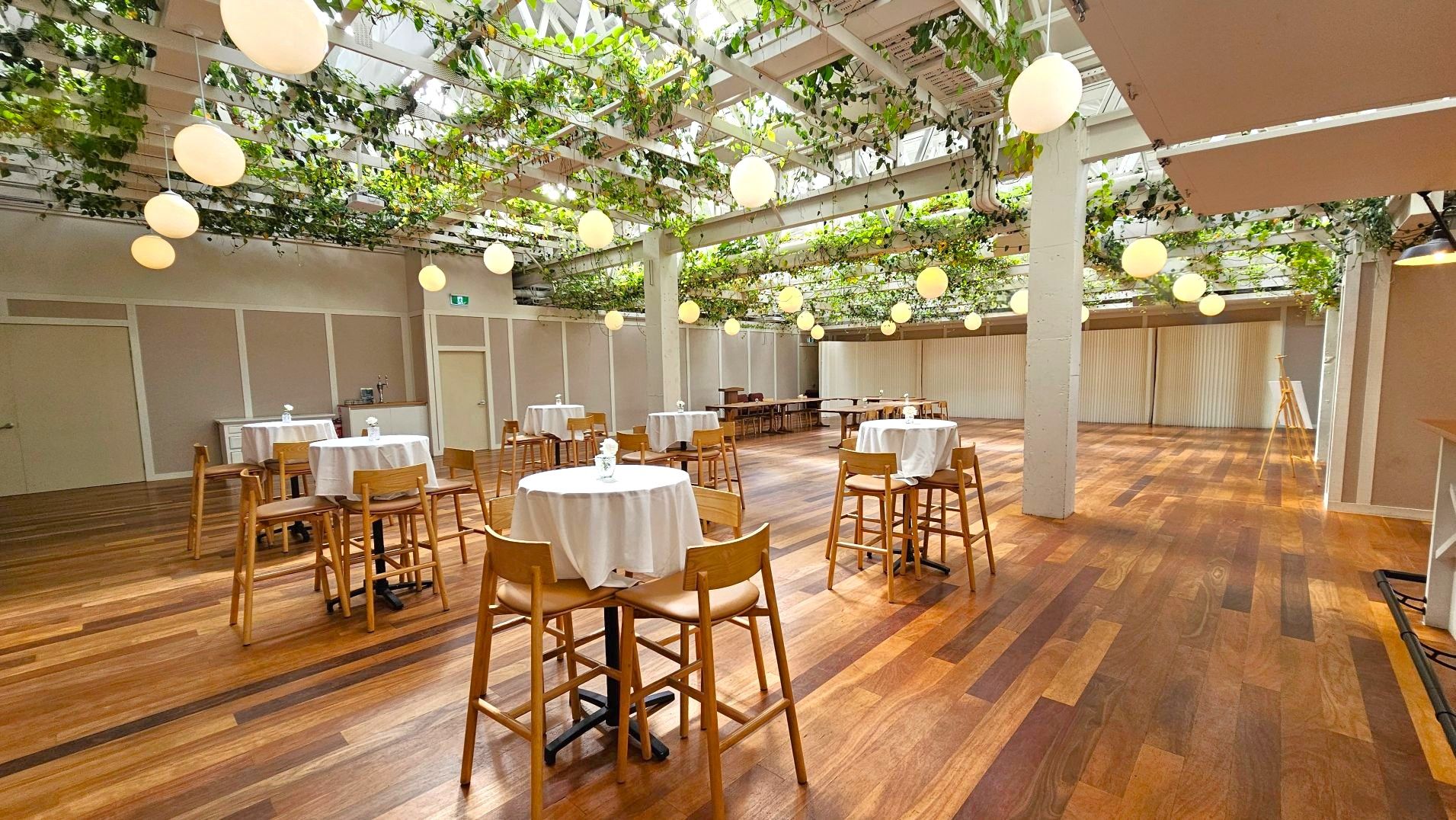 A spacious indoor venue with wooden flooring and a ceiling adorned with hanging greenery and round pendant lights. The room features several tall tables covered with white tablecloths, each surrounded by wooden chairs. A long counter is visible along one wall, and the walls are lined with light-coloured panels. The space is open and airy, with a modern, natural aesthetic.