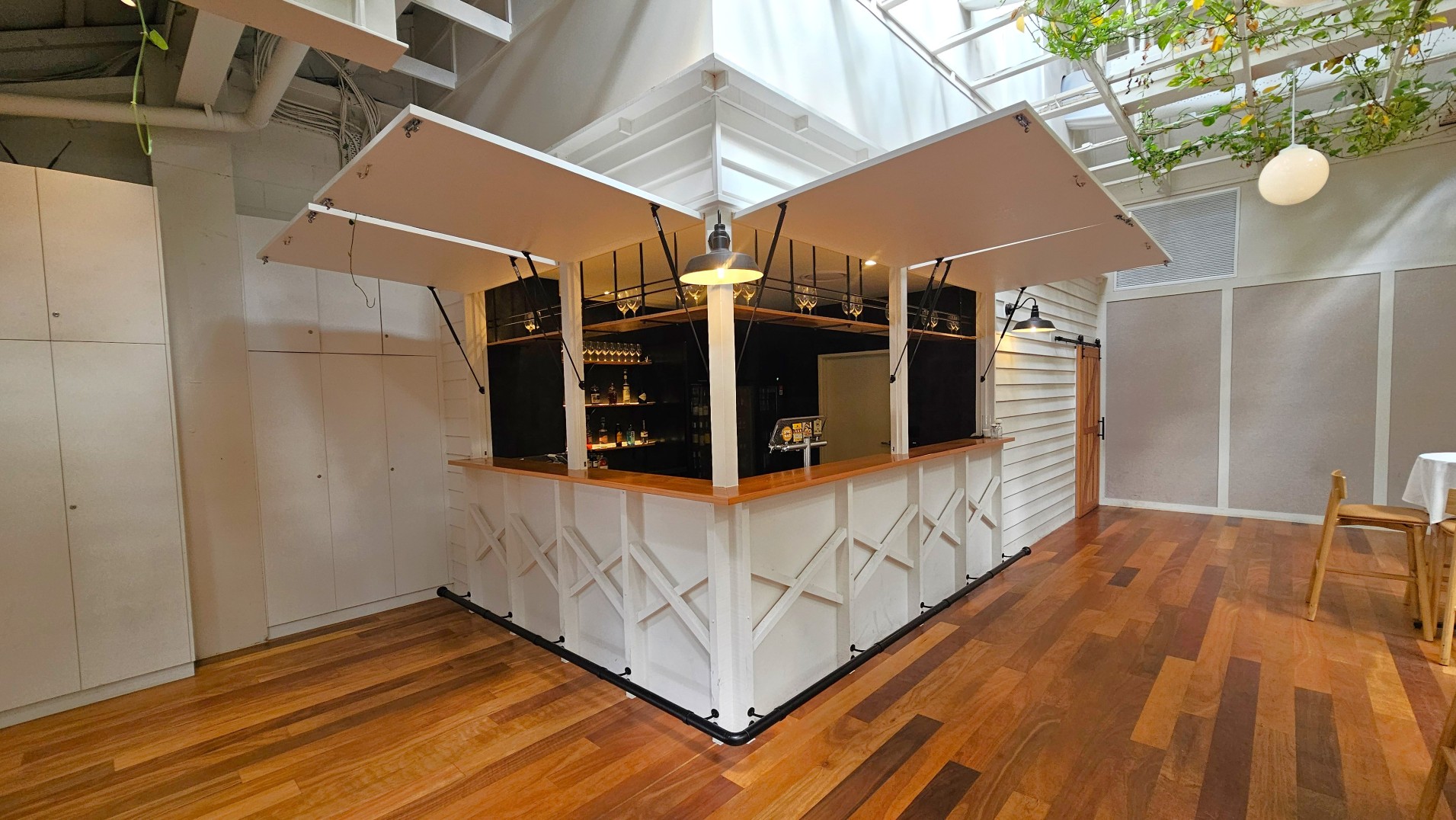 A small, white, kiosk-style bar with wooden countertops is situated in a corner of a room with wooden flooring. The bar has open panels revealing shelves with bottles and glasses. Above, a metal light fixture hangs centrally. The walls are white with a sliding wooden door on the right. Overhead, a glass ceiling with green plants adds a touch of nature. A single chair and table are partially visible on the right.