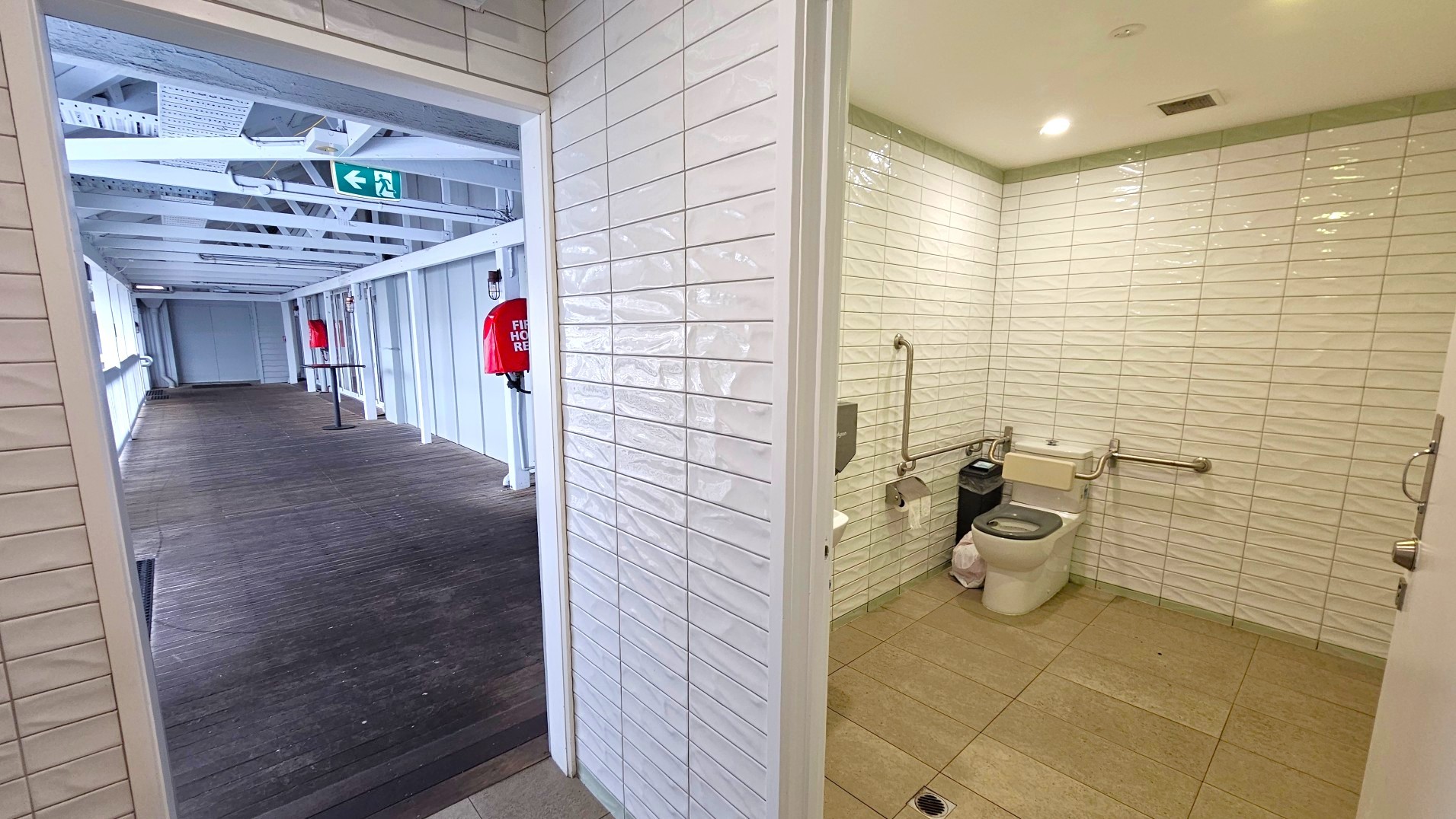 The image shows a corridor leading to a bathroom. The corridor has wooden flooring and white walls with exposed beams on the ceiling. A green emergency exit sign is visible above. The bathroom, visible through an open door, has white tiled walls and a beige tiled floor. It features a toilet with grab bars, a paper dispenser, and a waste bin. The space is well-lit with recessed ceiling lights.