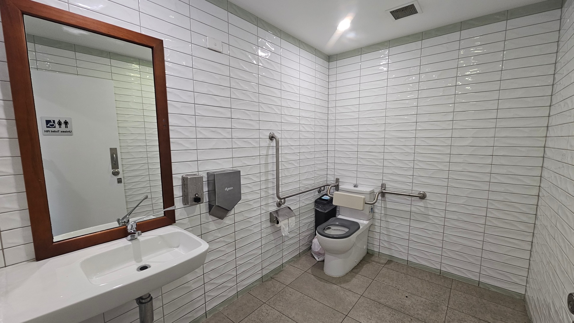 A clean, accessible toilet with white tiled walls and a beige tiled floor. A white sink with a mirror above is on the left, accompanied by a soap dispenser and a hand dryer. The toilet is on the right, equipped with grab bars on both sides and a green seat cover. A waste bin is nearby. The door, visible in the mirror, has a sign indicating a unisex toilet with wheelchair access.
