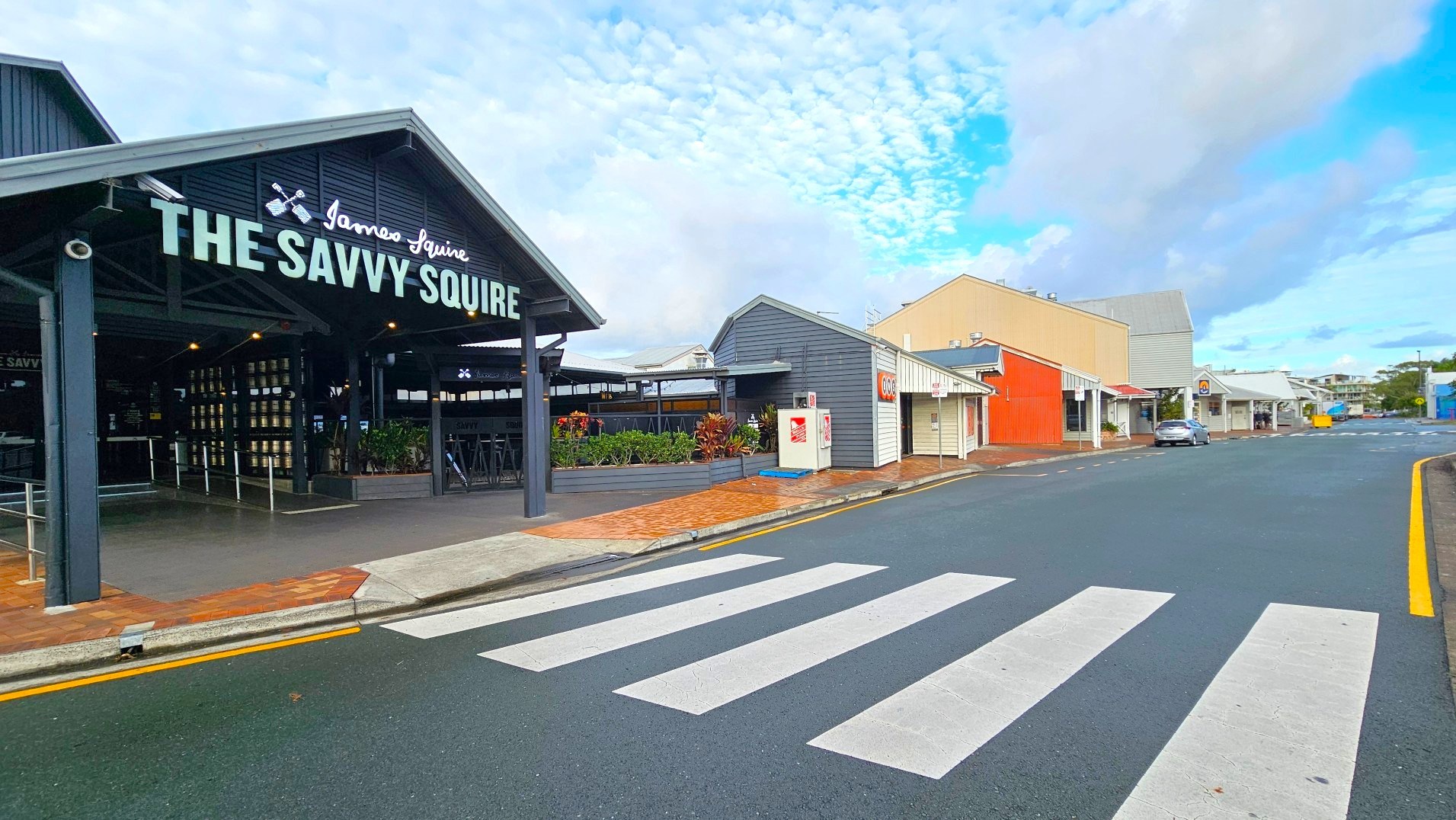A street view featuring a prominent pub with a large sign reading 'The Savvy Squire' in bold letters. The pub has a dark exterior with open seating visible inside. Adjacent buildings are small, with varied colours including grey and red. A zebra crossing leads to the pub entrance, and the road is bordered by a yellow line. The area appears open with a mix of brick and concrete surfaces.