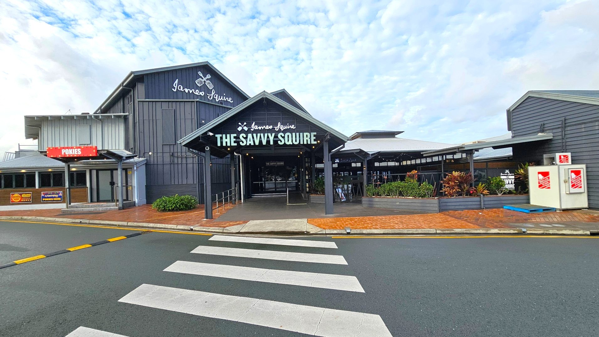 A modern building with a prominent entrance labelled 'The Savvy Squire' in bold letters. The structure features a mix of dark and light grey cladding with a pitched roof. To the left, a sign reads 'POKIES' above a smaller entrance. A zebra crossing leads to the main entrance, flanked by planters with greenery. The area is paved with red bricks, and a small ramp provides access to the entrance.
