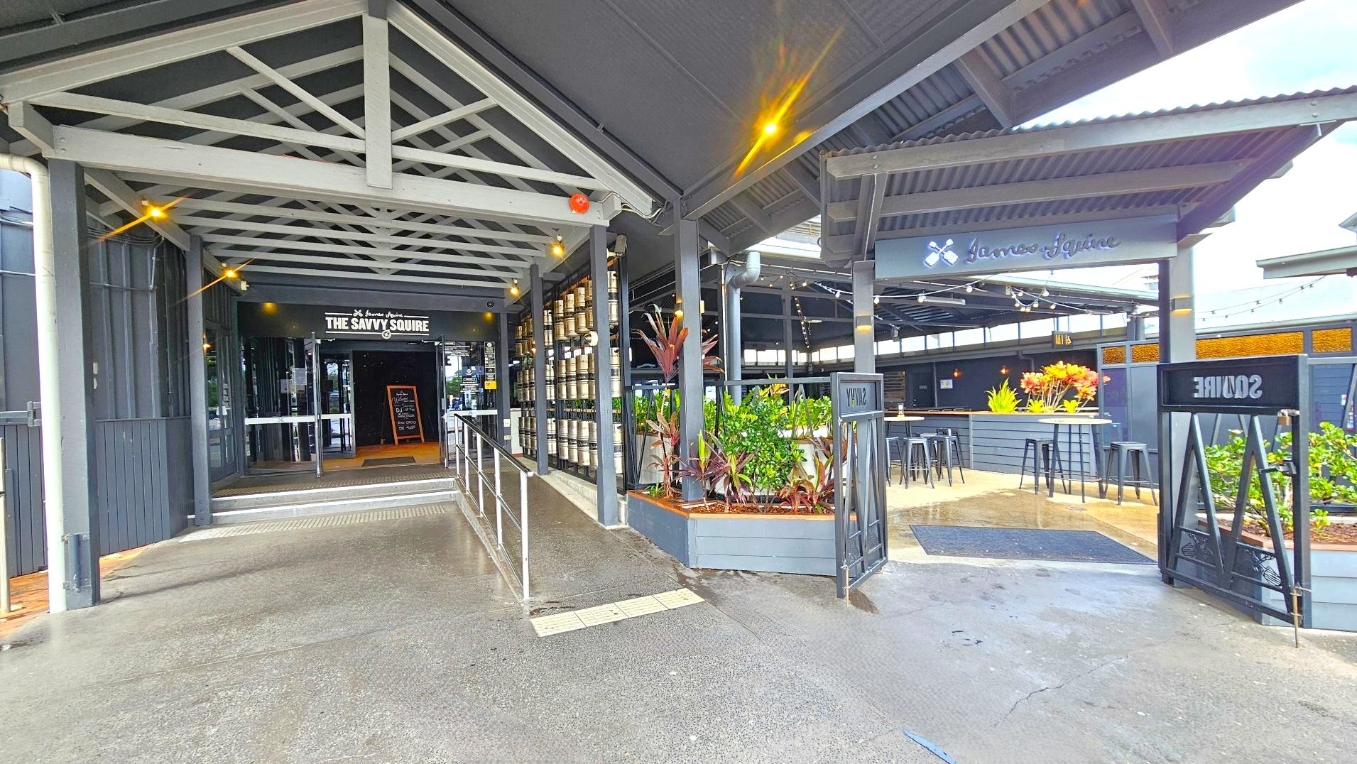 The image shows an entrance to a venue named 'The Savvy Squire'. The entrance features a covered walkway with a metal truss roof and a ramp with handrails leading to glass doors. To the right, there's an open seating area with high tables and stools, surrounded by planters with greenery. The flooring transitions from concrete to a textured surface at the entrance. Signage is visible above the entrance and seating area.