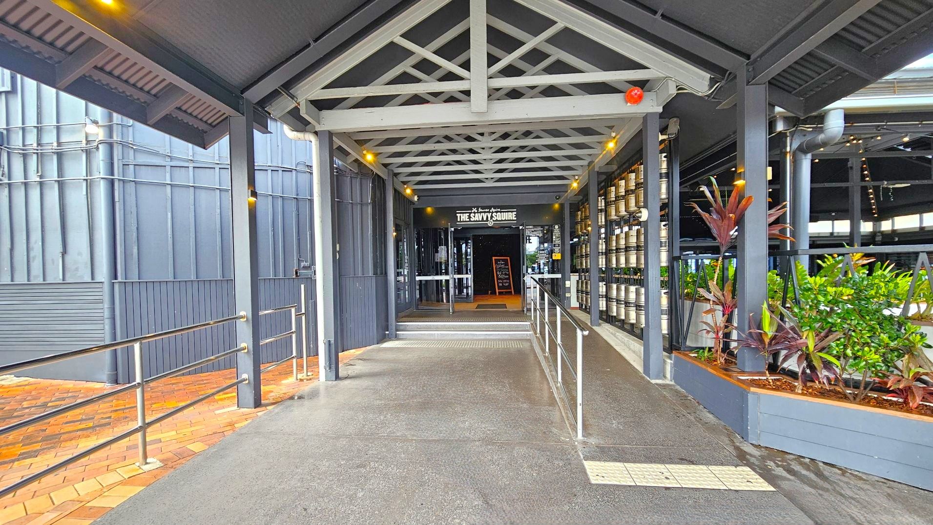 A covered walkway leads to the entrance of a venue named 'The Savvy Squire'. The path is flanked by metal railings and bordered by a row of plants on the right. The floor transitions from concrete to brick near the entrance. The structure features a metal roof with exposed beams and is enclosed by walls on the left. A blackboard sign is visible near the glass doors at the entrance.