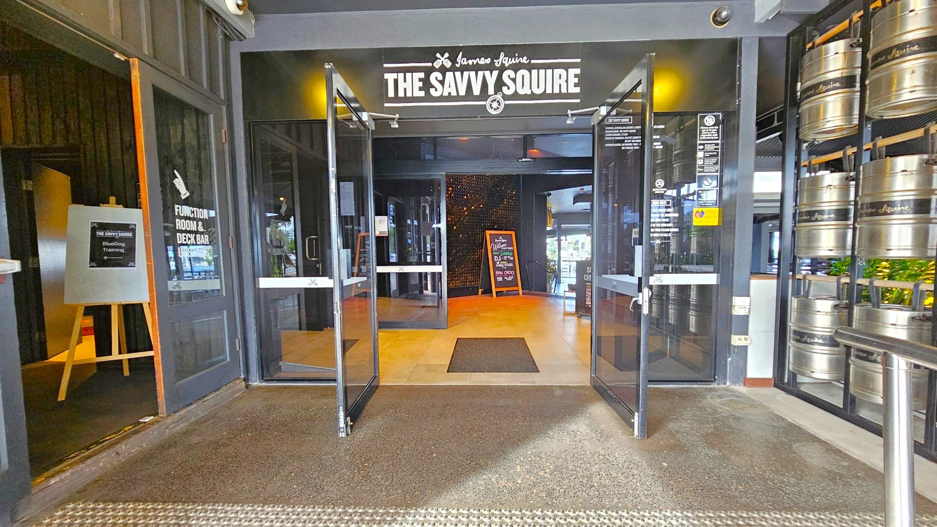 The entrance to 'The Savvy Squire' features two open glass doors with metal frames, leading into a warmly lit interior. A sign above the doors displays the venue's name. To the left, a sign on an easel reads 'BlueDog Training'. On the right, metal kegs are stacked neatly. The floor transitions from textured concrete outside to smooth tiles inside. A blackboard with event details stands inside, enhancing the welcoming atmosphere.