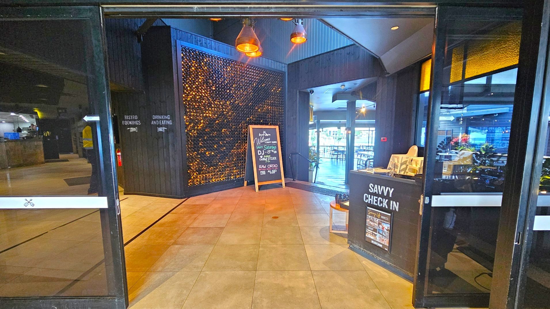 The image shows an entrance area with a tiled floor leading into a venue. On the left, a wall with a decorative display of wine bottles is visible. A chalkboard sign stands in front, advertising events. To the right, a counter labelled 'Savvy Check In' is present, with brochures and a high chair. The space is enclosed by glass doors and dark walls, with pendant lights hanging from the ceiling.