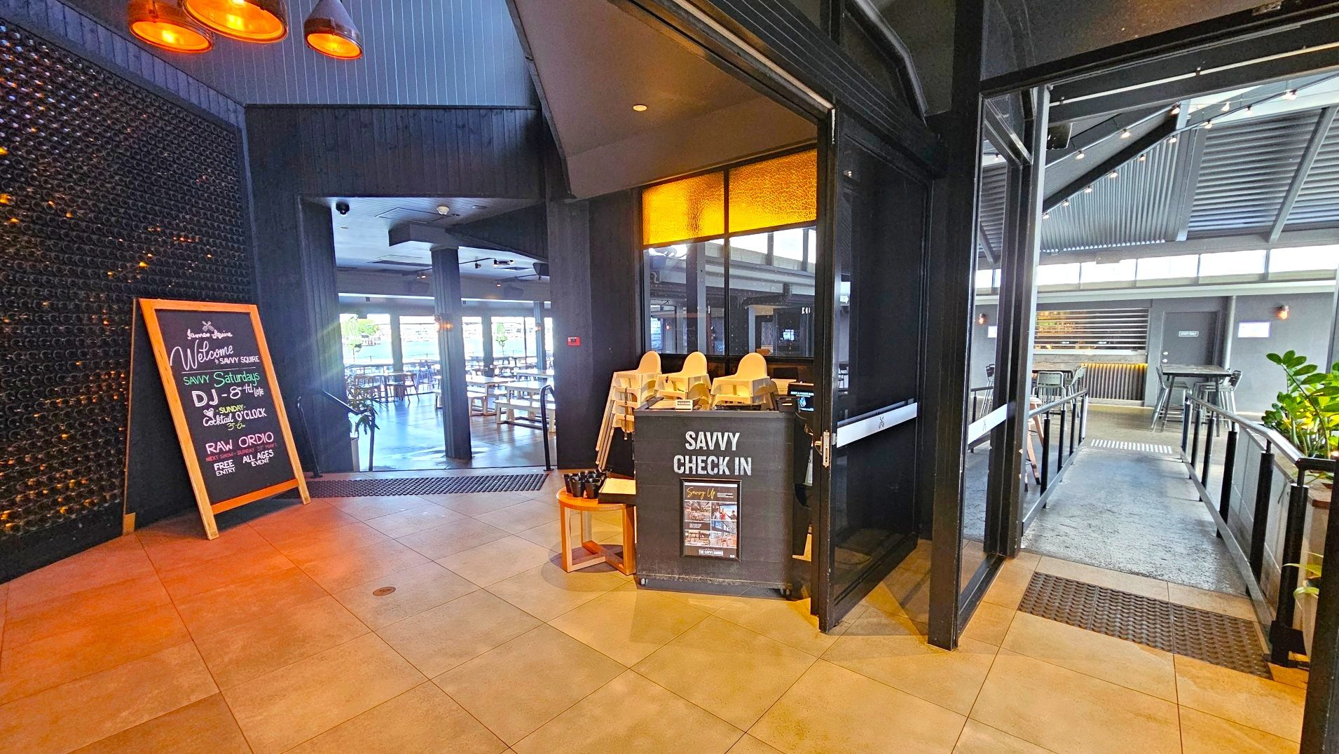 The image shows an entrance area with a tiled floor leading into a restaurant or bar. A large blackboard sign on the left welcomes guests and lists events. To the right, a 'Savvy Check In' station with high chairs is positioned near a glass door. Beyond, a ramp with metal railings leads to an open seating area with tables and chairs. The space is enclosed by dark walls and a corrugated metal ceiling.
