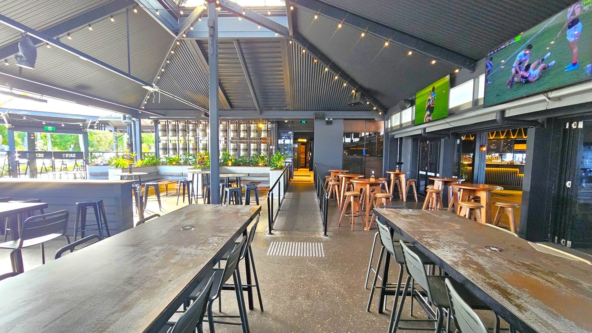 The image shows a spacious indoor bar area with a high, corrugated metal ceiling and string lights. Long wooden tables with black chairs are arranged in rows. To the right, there are high wooden stools and tables. Large screens displaying sports are mounted on the wall. A row of beer kegs and potted plants line the back. The floor is polished concrete, and a ramp with railings leads to a doorway at the back.