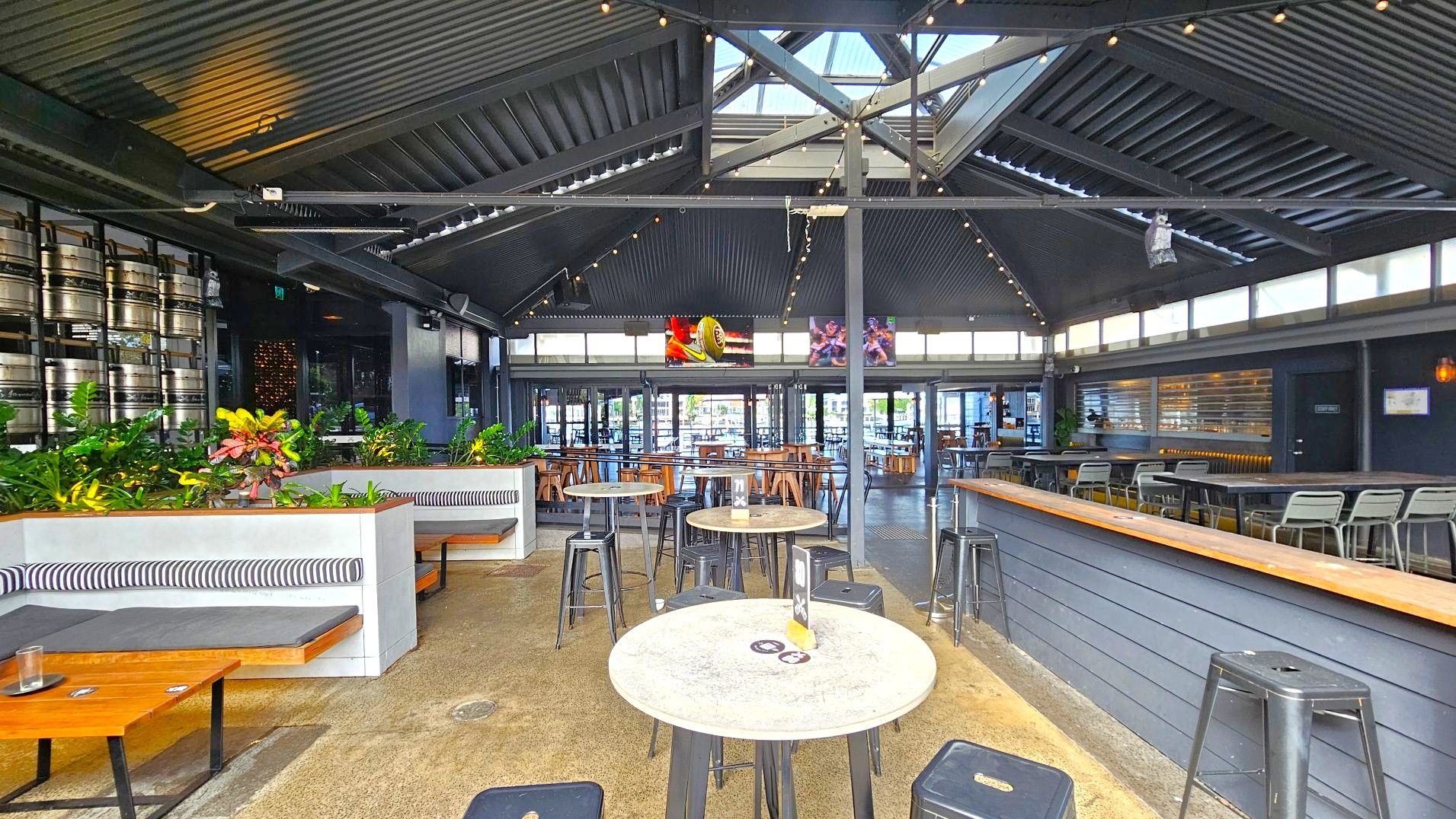The interior of a modern bar with a high, dark corrugated metal ceiling featuring skylights. The space includes a mix of seating options: cushioned booths with striped backs, round tables with metal stools, and a long bar with high chairs. Green plants add a touch of nature. Large windows and glass doors line the far wall, allowing natural light. Kegs are stacked on one side, and two screens display sports images.