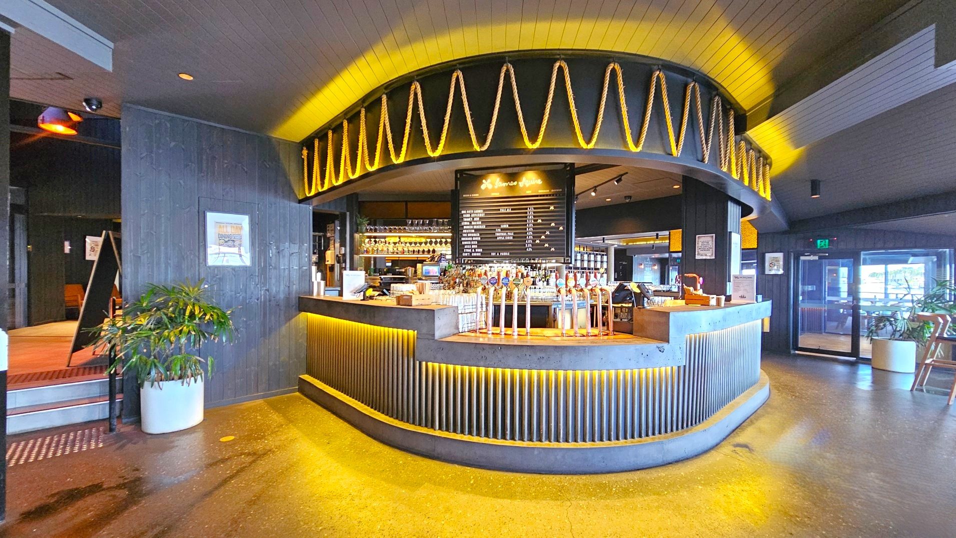 A modern bar area with a curved counter made of concrete and wood, featuring a row of beer taps. Above the counter, a decorative rope design is illuminated with warm lighting. Behind the bar, shelves display glasses and bottles, and a menu board lists drink options. The floor is polished concrete, and there are potted plants on either side. An exit door with glass panels is visible to the right.