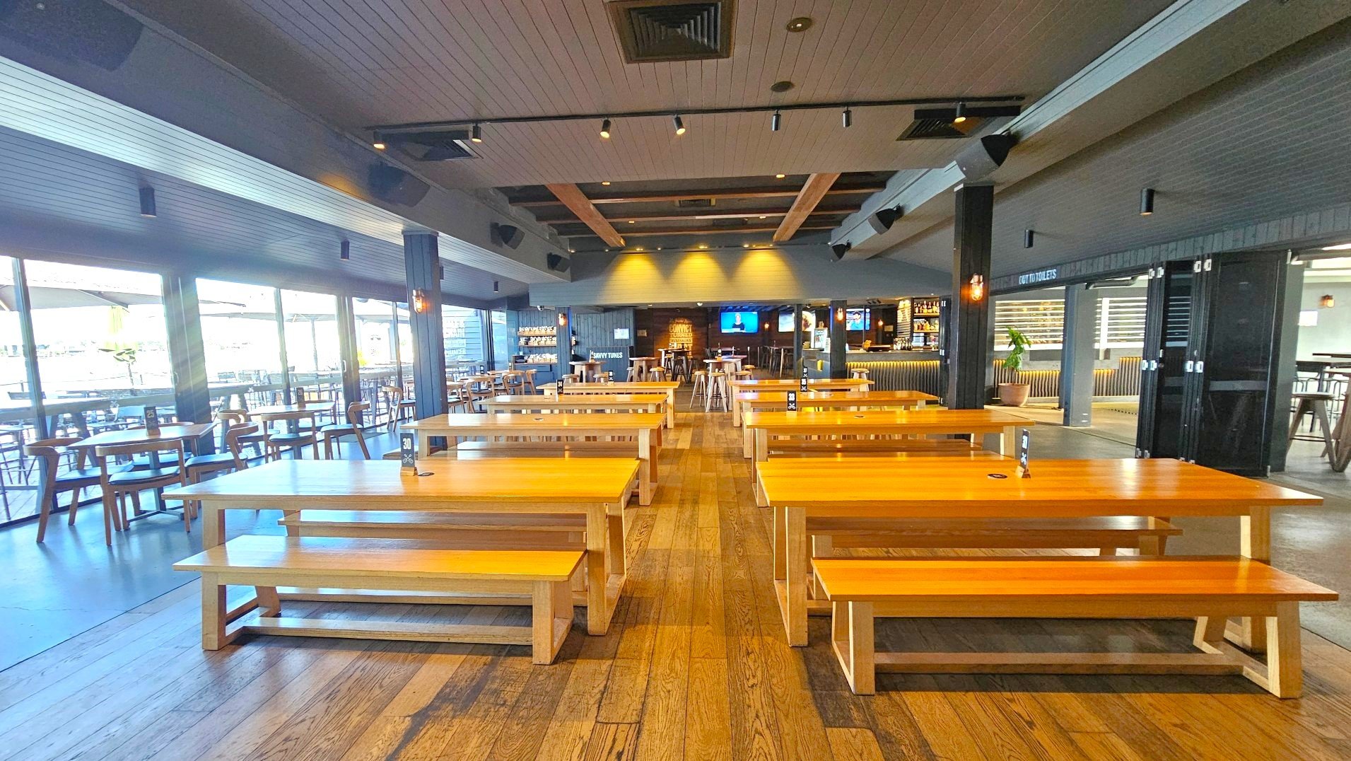 A spacious indoor dining area with wooden tables and benches arranged in rows on a wooden floor. The ceiling features exposed beams and recessed lighting. Large glass doors on the left open to an outdoor seating area. A bar with stools is visible at the back, along with a wall-mounted TV. A sign indicating toilets is on the right. The space feels open with a mix of natural and artificial lighting.
