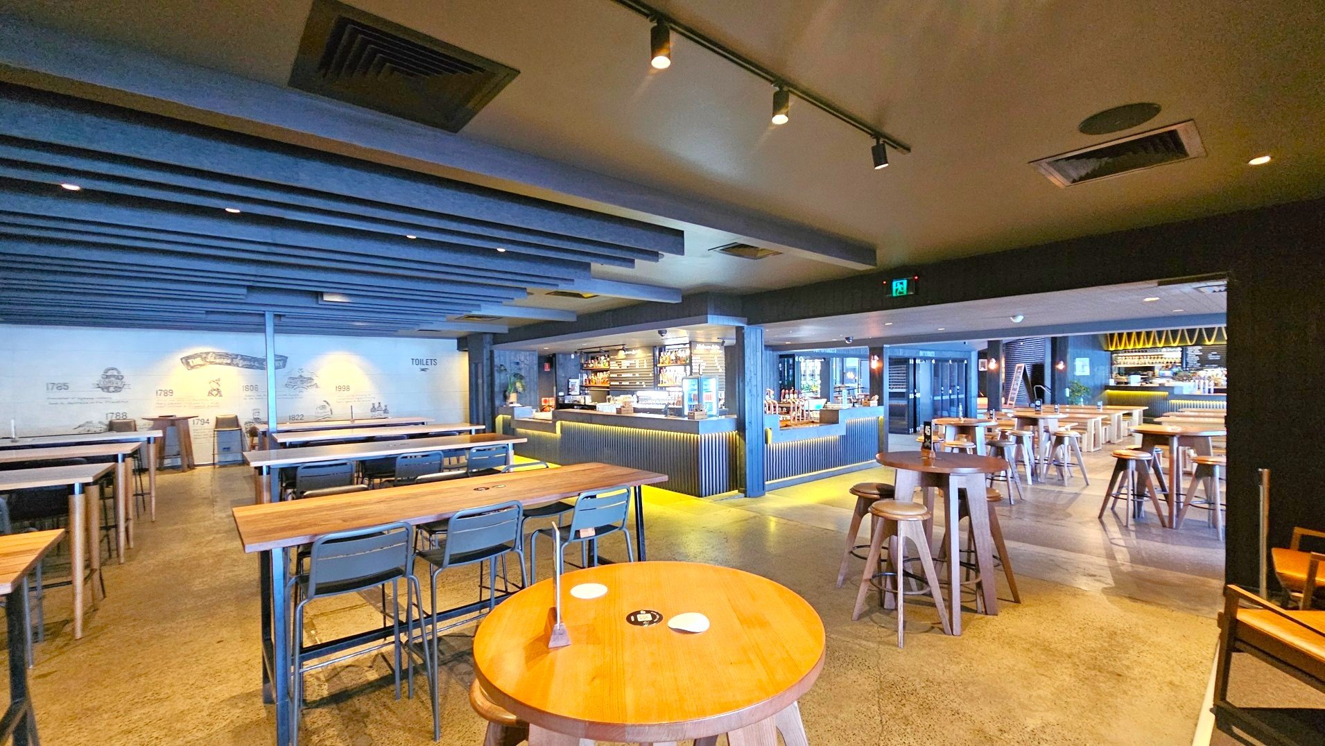 The image shows a spacious indoor dining area with a modern design. Wooden tables and high stools are arranged neatly across the polished concrete floor. A prominent bar counter with a corrugated metal front is visible, stocked with drinks and snacks. The ceiling features exposed beams and spotlights. A wall with historical illustrations and a 'Toilets' sign is on the left. The space feels open and inviting.