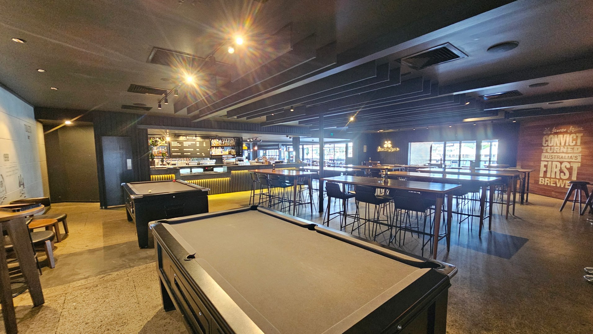 The image shows a spacious bar interior with a modern design. In the foreground, there are two pool tables with dark surfaces. To the right, high tables with bar stools are arranged in rows. The bar counter is visible in the background, illuminated with warm lighting and stocked with bottles. The floor is a mix of polished concrete and wood. A large wall sign reads 'Convict Turned Australia's First Brewer'.