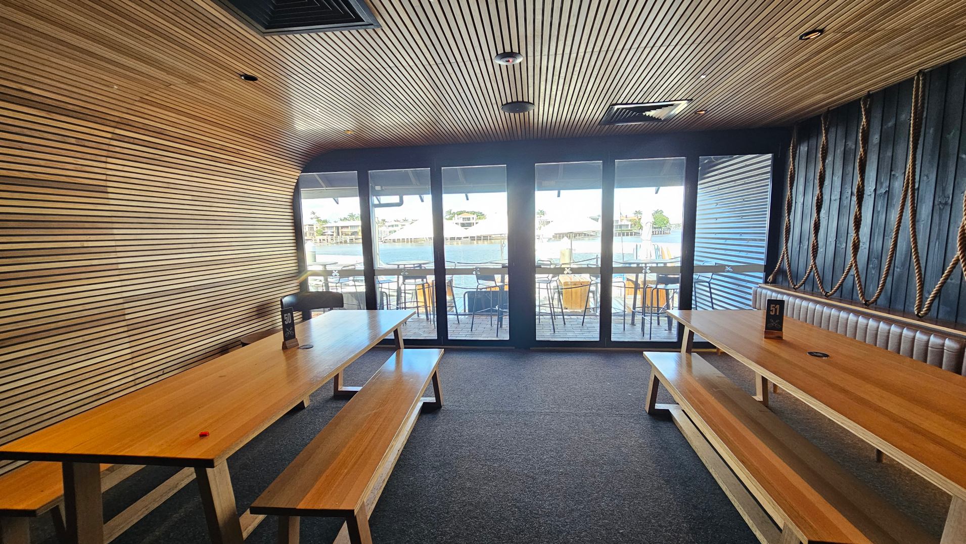 The interior of a room with wooden slatted walls and ceiling, featuring two long wooden tables with benches on either side. The right wall is adorned with thick ropes. Large glass doors at the far end open to an outdoor seating area with views of water and buildings. The floor is carpeted in a dark hue, and table numbers are visible on the tables.