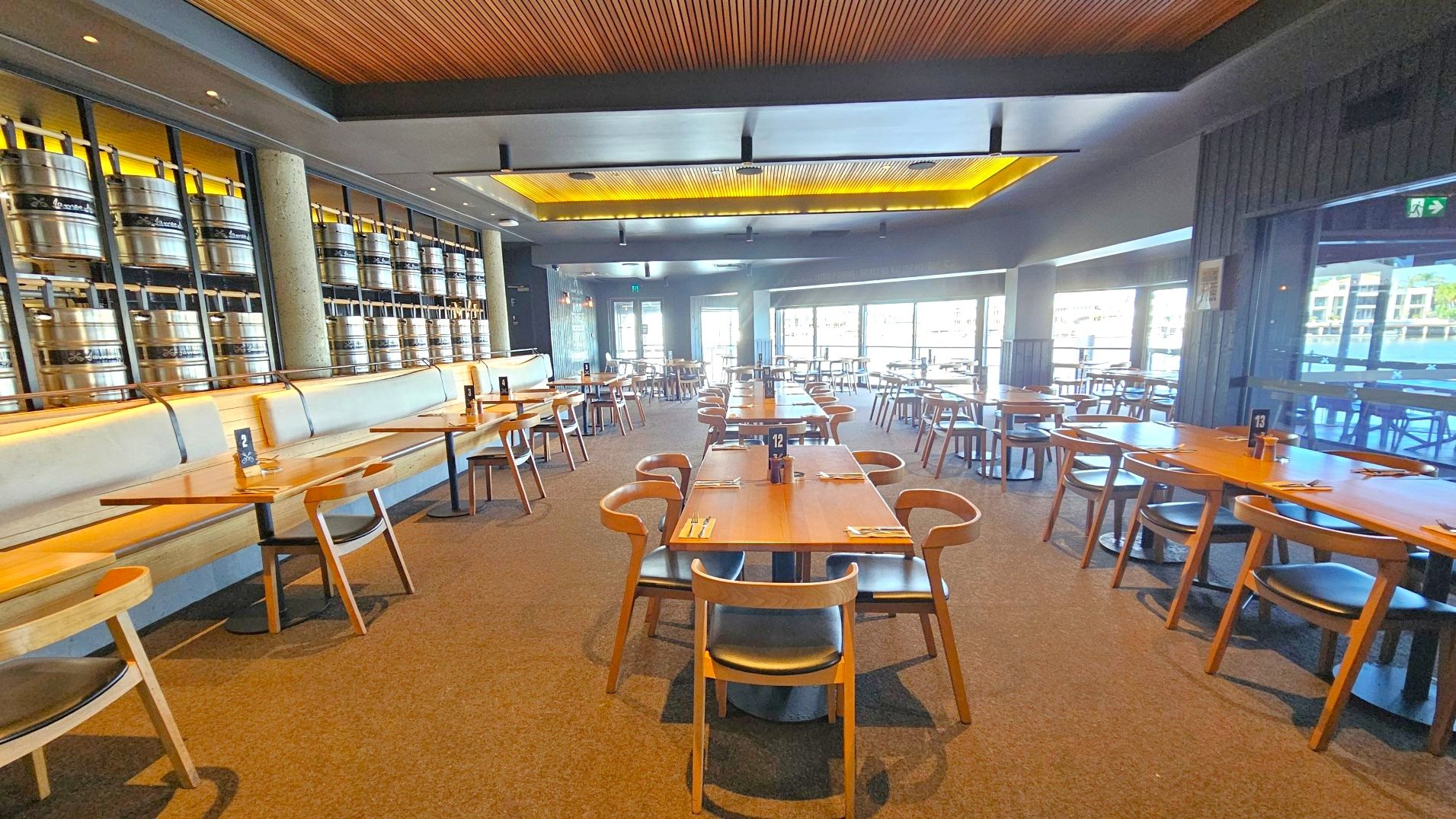 A spacious restaurant interior with rows of wooden tables and chairs arranged neatly on a carpeted floor. The left wall features a series of large metal barrels stacked in a display, with a long cushioned bench running parallel. The ceiling has a wooden panel design with recessed lighting. Large windows on the right allow natural light to flood in, offering views of a waterfront. Numbered table signs and cutlery are set on each table.