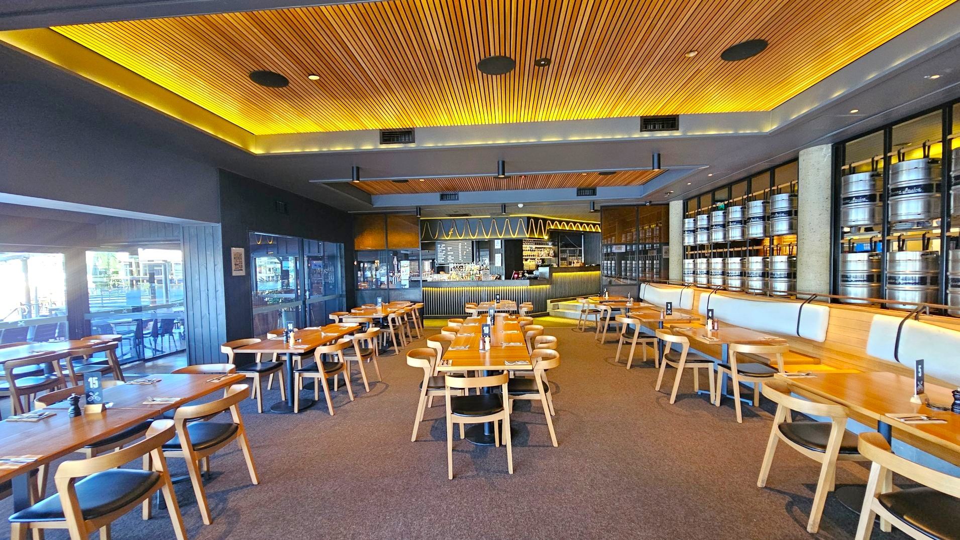 A modern restaurant interior featuring wooden tables and chairs arranged neatly on a carpeted floor. The ceiling is lined with wooden slats and recessed lighting, creating a warm ambience. A bar with a zigzag light pattern is visible at the back, flanked by shelves of metal kegs. Large windows on the left allow natural light in, and a long cushioned bench lines the right wall.