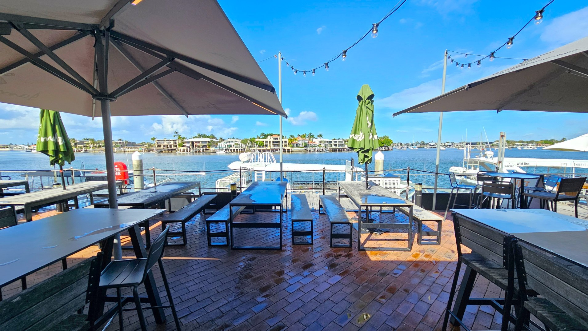 An outdoor dining area with several rectangular tables and benches on a red brick patio. Large umbrellas provide shade, and string lights are suspended above. The area overlooks a body of water with boats docked nearby. Across the water, there are residential buildings. A railing separates the dining area from the water, and a lifebuoy is attached to it. The scene is bright and open, with a clear view of the surroundings.