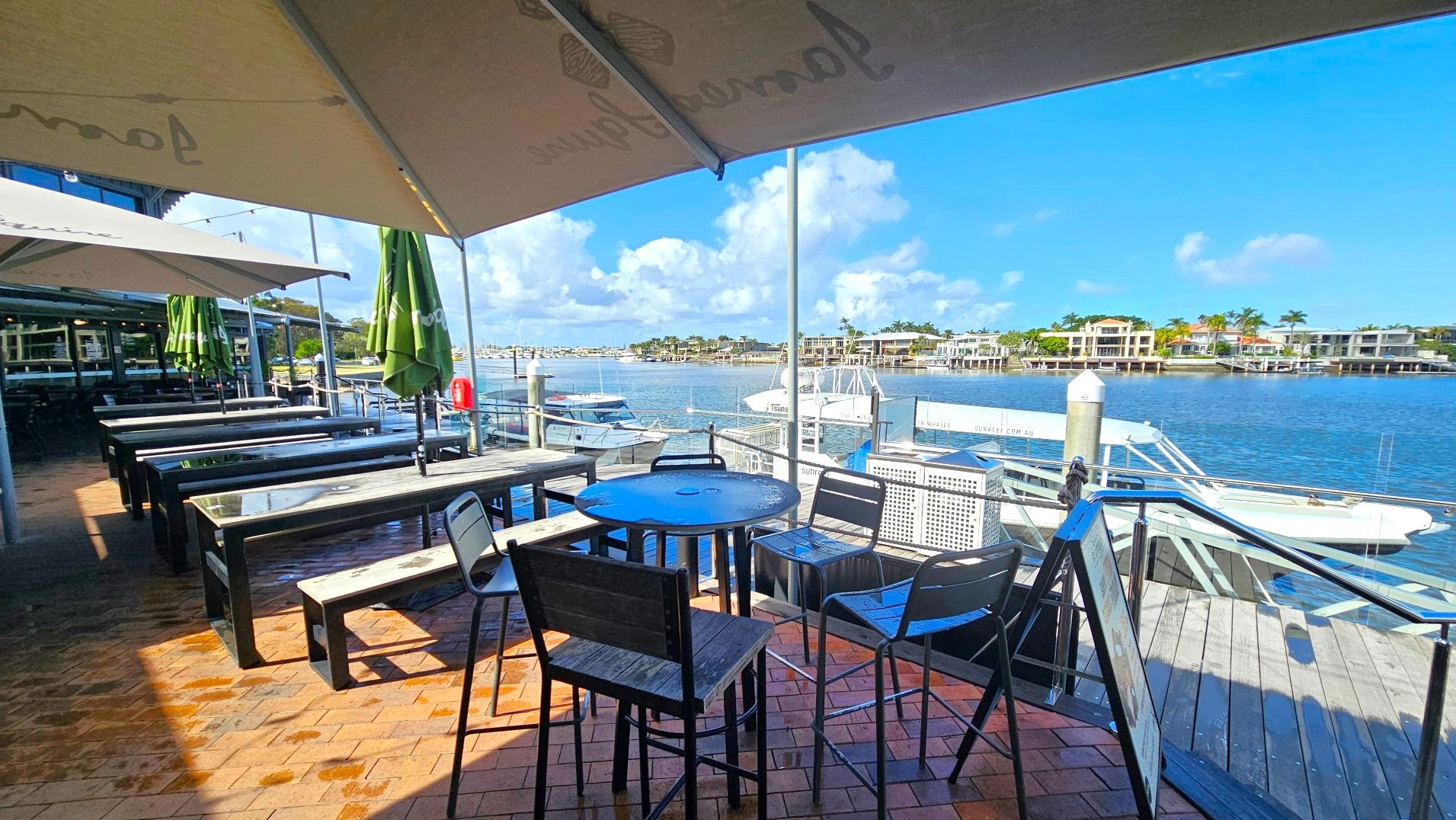 An outdoor dining area with wooden tables and chairs is set on a brick patio under large umbrellas. The area overlooks a marina with several boats docked along the water. Glass railings separate the dining space from the water. In the background, there are waterfront houses with palm trees. The scene is bright and open, with a clear view of the marina and surrounding buildings.