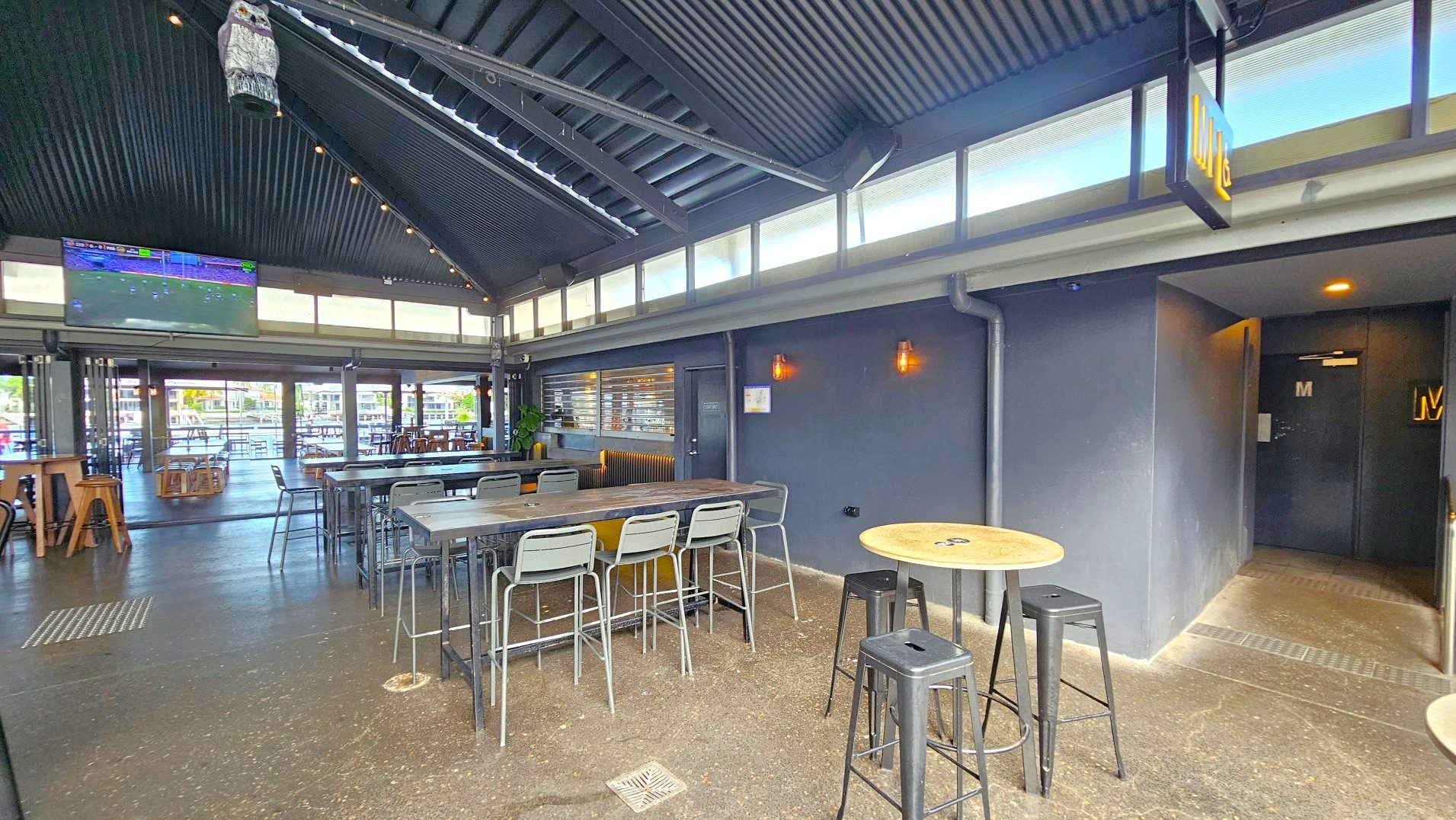 The interior of a modern bar with a high, corrugated metal ceiling and large windows near the roof allowing natural light. A large TV screen is mounted on the wall showing a sports game. The space features high tables with metal stools and a polished concrete floor. A sign marked 'M' indicates a restroom entrance on the right. The area feels open with visible seating extending outside.