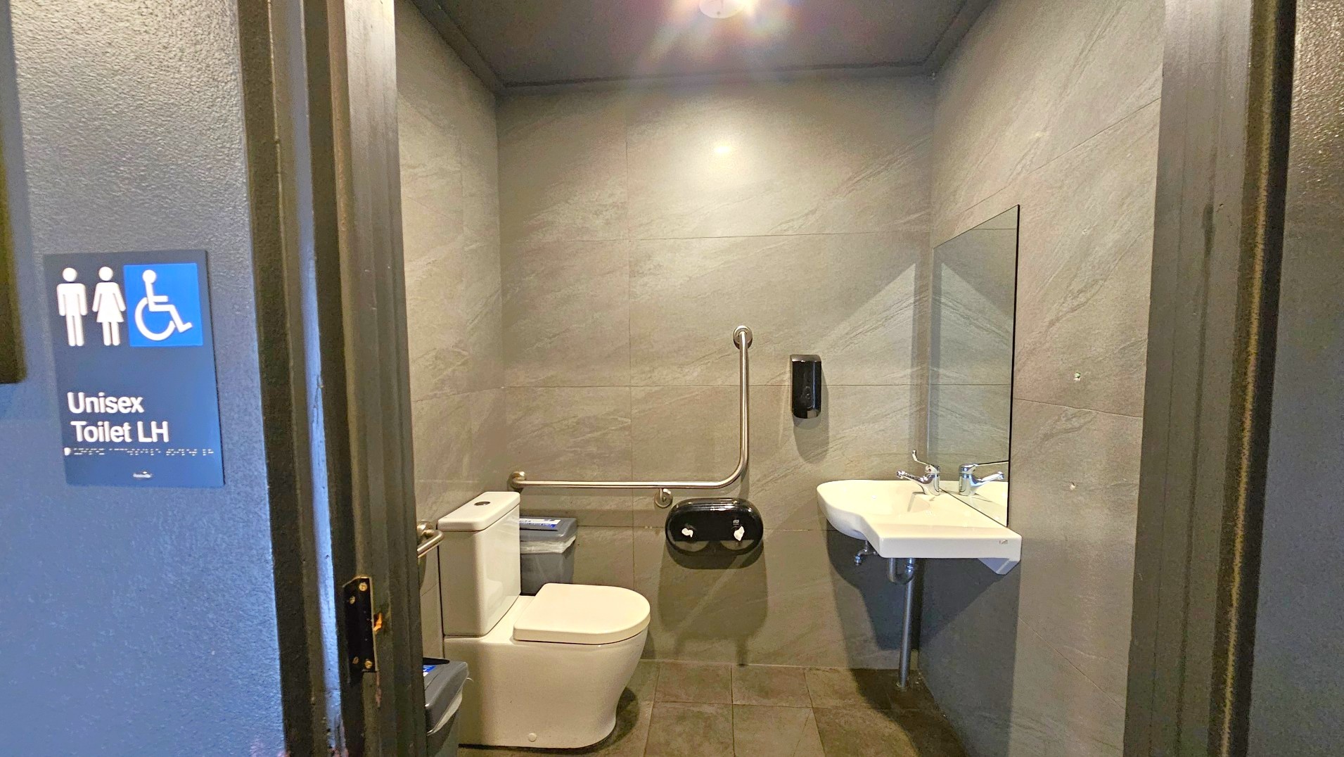 A unisex accessible toilet with grey tiled walls and floor. The room features a white toilet with a silver grab rail on the wall beside it. A black toilet paper dispenser and a soap dispenser are mounted on the wall. A white sink with a mirror above it is on the right. The entrance door is open, displaying a sign with male, female, and wheelchair symbols, labelled 'Unisex Toilet LH'.