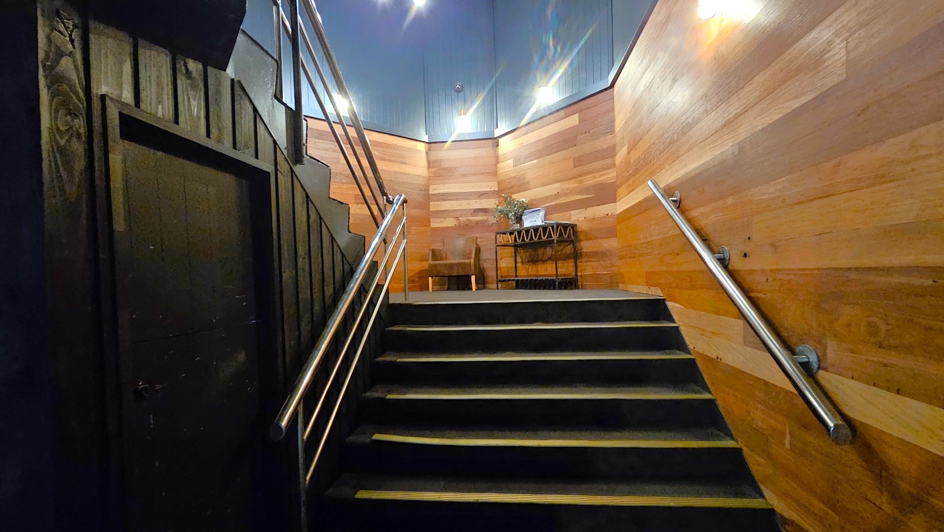 A staircase with dark steps leads up to a small landing area. The walls are clad in horizontal wooden panels, creating a warm, enclosed feel. Stainless steel handrails are mounted on both sides of the stairs. At the top, a wooden table with a decorative plant and a chair are visible. The ceiling is painted a dark colour, and several wall-mounted lights illuminate the space.