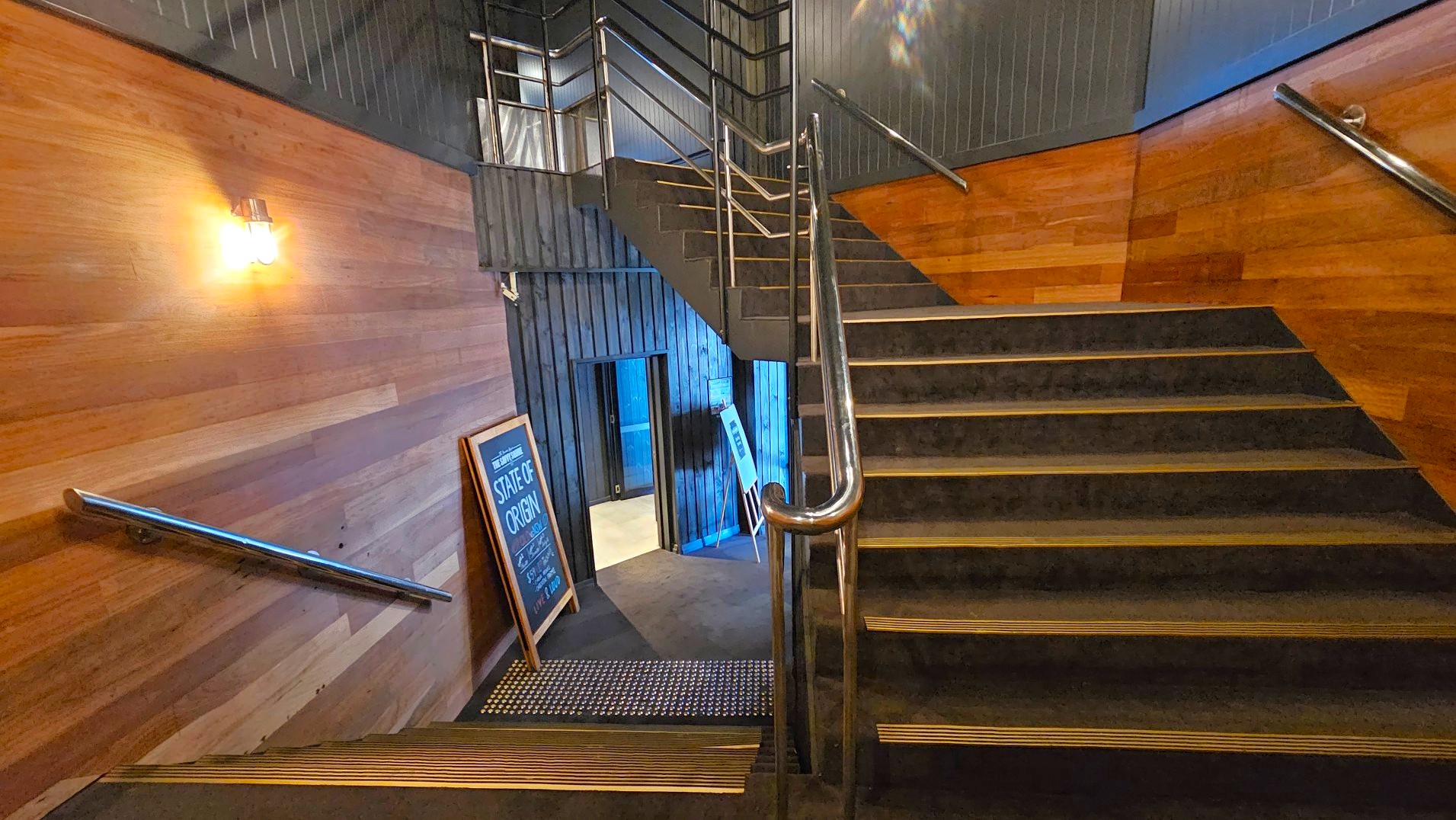 A staircase with wooden panelling on the walls and metal railings leads up and down. The stairs are carpeted with black treads and yellow safety edges. A lit wall sconce illuminates the area. At the bottom, a chalkboard sign reads 'State of Origin'. The space feels enclosed with dark wood and metal accents. A small doorway is visible at the bottom, leading to another area.