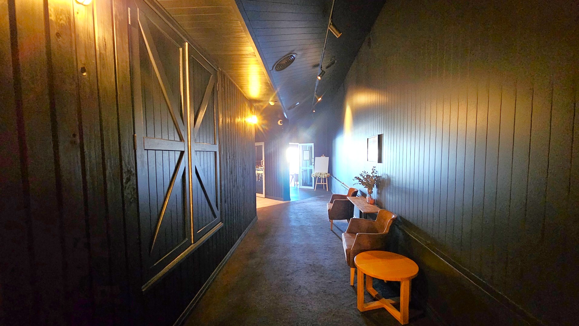 A dimly lit corridor with dark wooden panelled walls and ceiling, featuring a large barn-style sliding door on the left. On the right, a small seating area with two brown chairs and a round wooden table is visible, along with a narrow console table holding a vase with foliage. The carpeted floor leads to a brighter area at the end of the corridor, where a doorway opens into another room.