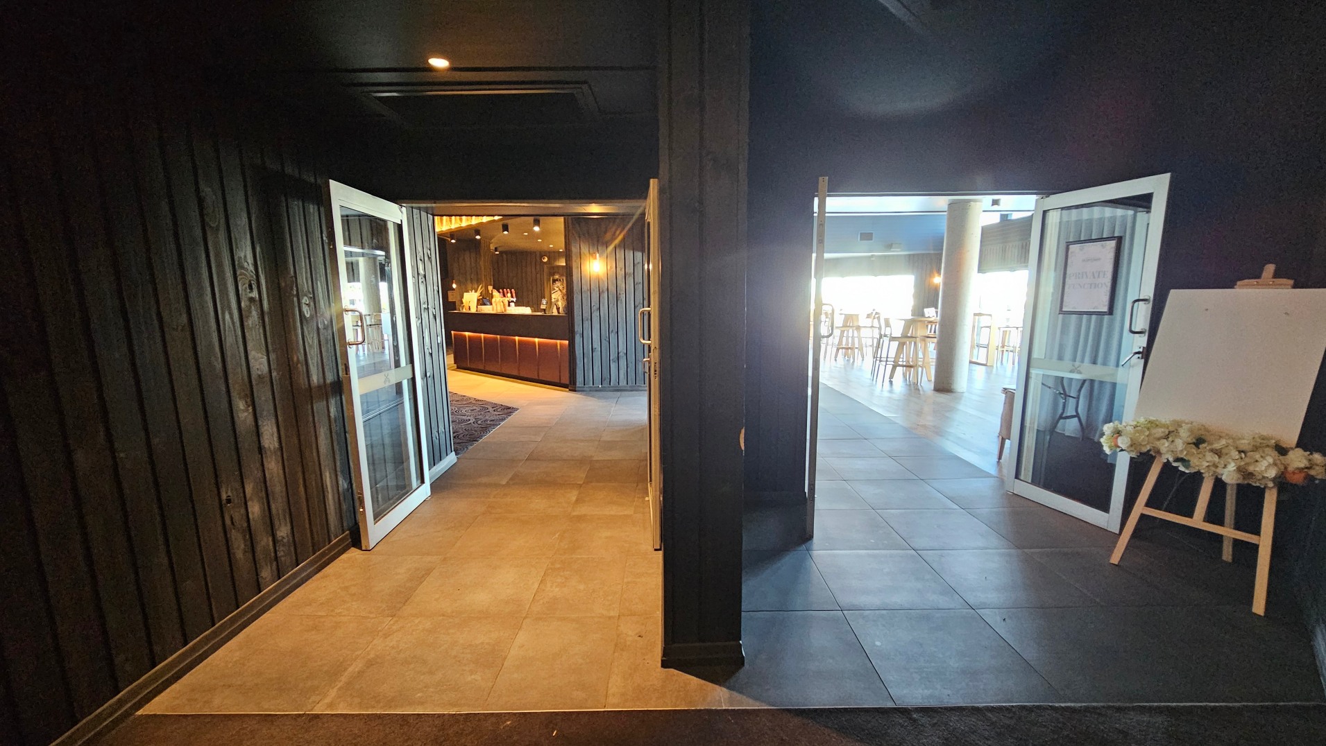 The image shows an interior space with two open doorways leading to different areas. On the left, a doorway opens to a warmly lit bar area with wooden panelling and a counter. On the right, another doorway leads to a brighter space with high tables and stools, featuring a large window. The floor transitions from dark tiles to lighter tiles. An easel with floral decorations stands near the right doorway.