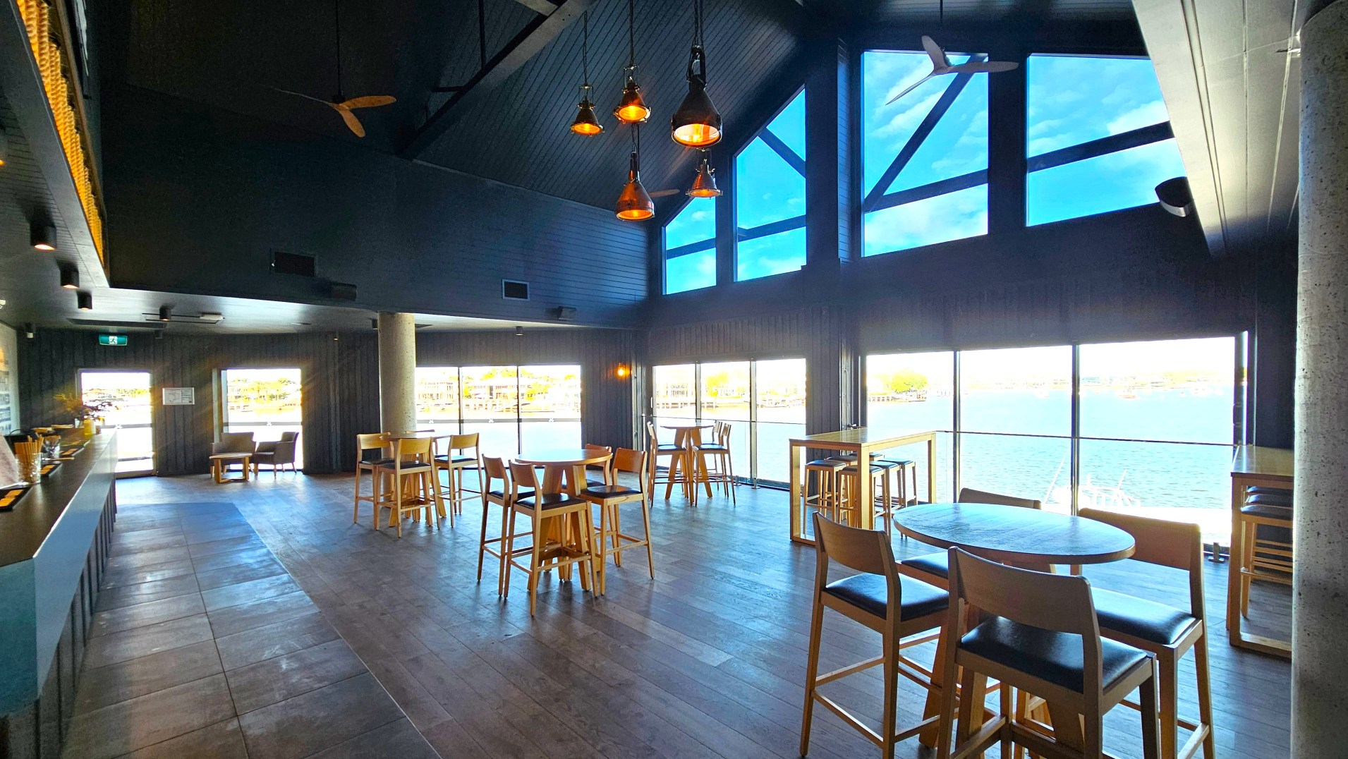 The interior of a modern, open-plan restaurant with large floor-to-ceiling windows offering a view of a waterfront. The space features dark wooden floors and ceiling, with several wooden tables and chairs arranged throughout. Pendant lights hang from the ceiling, and a long counter is visible on the left. A concrete pillar stands near the centre, and the room feels spacious and airy due to the high ceiling and expansive windows.