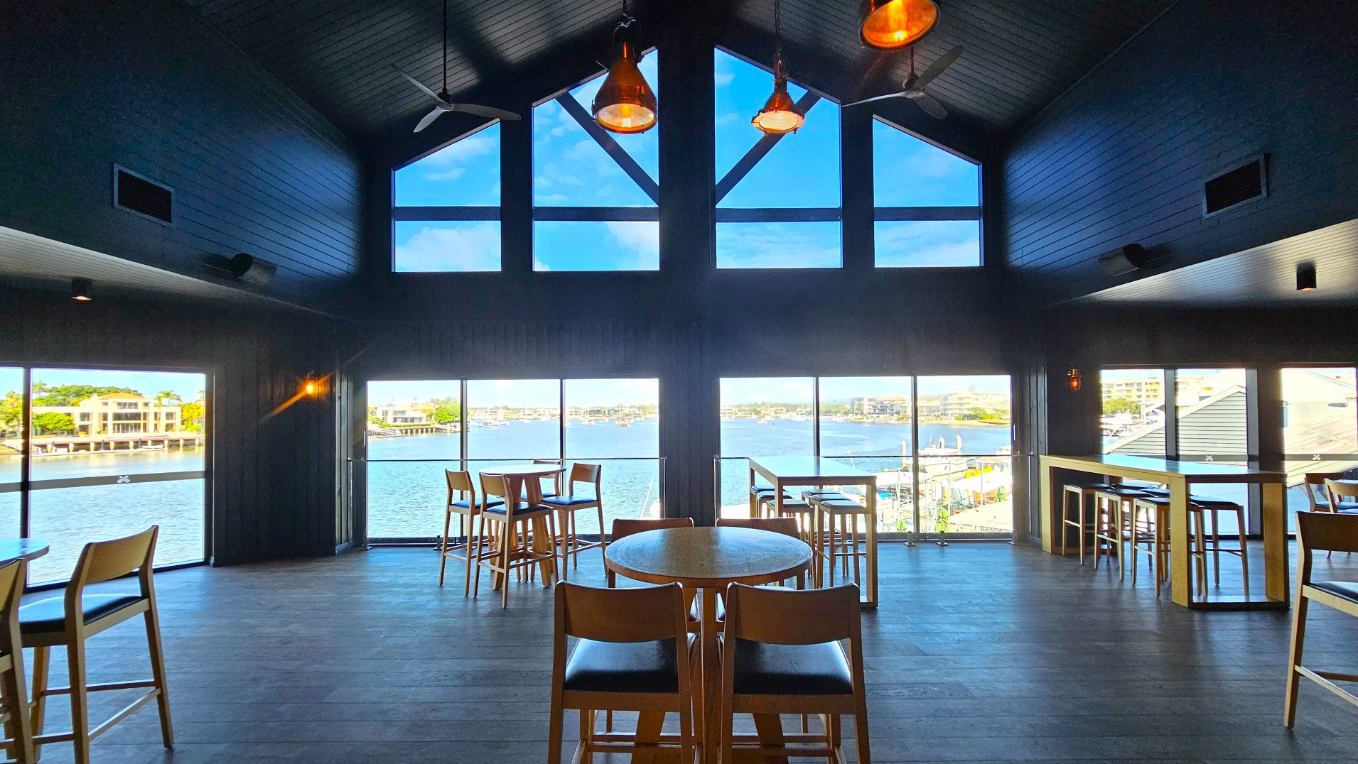 A spacious interior with large floor-to-ceiling windows offering a view of a waterfront with buildings and boats. The room features dark wooden floors and a high, vaulted ceiling with exposed beams. Several wooden tables and chairs are arranged throughout, with pendant lights hanging from the ceiling. The space feels open and airy, with a focus on the scenic view outside.