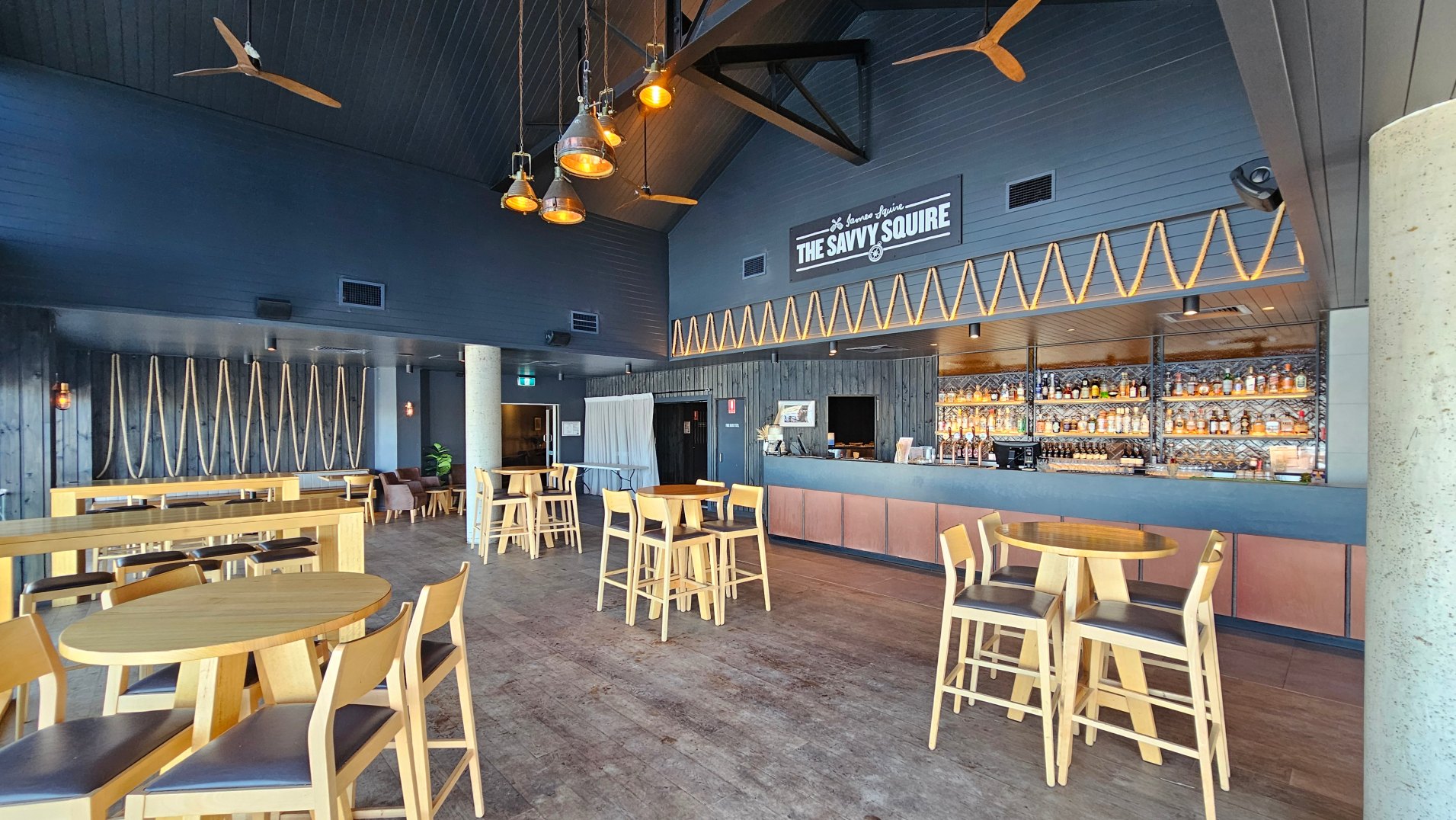 The interior of a modern bar with high ceilings and dark wood panelling. A prominent sign reads 'The Savvy Squire' above a well-stocked bar with illuminated shelves. Light wooden tables and chairs are scattered across a wooden floor. Decorative rope patterns adorn the walls, and pendant lights hang from the ceiling. A concrete pillar stands near the centre, and a small seating area is visible in the background.