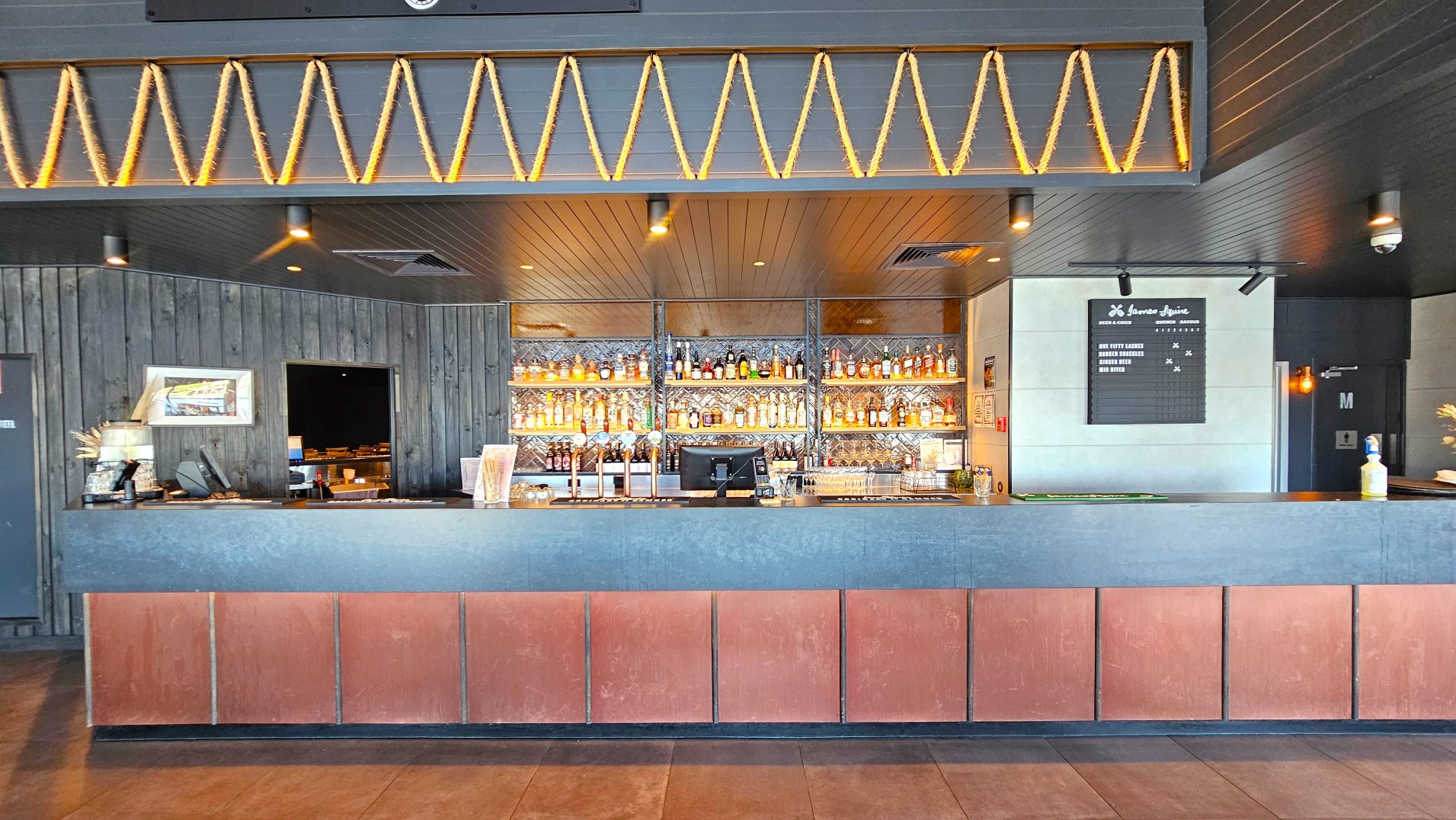 A bar area with a dark wood panelled wall and ceiling, featuring a prominent counter with a blue-grey top and reddish-brown front panels. Behind the counter, shelves display a variety of bottles, illuminated by warm lighting. A menu board is mounted on the right wall. The floor is tiled, and a decorative rope pattern adorns the upper wall. A doorway and a framed picture are visible on the left.