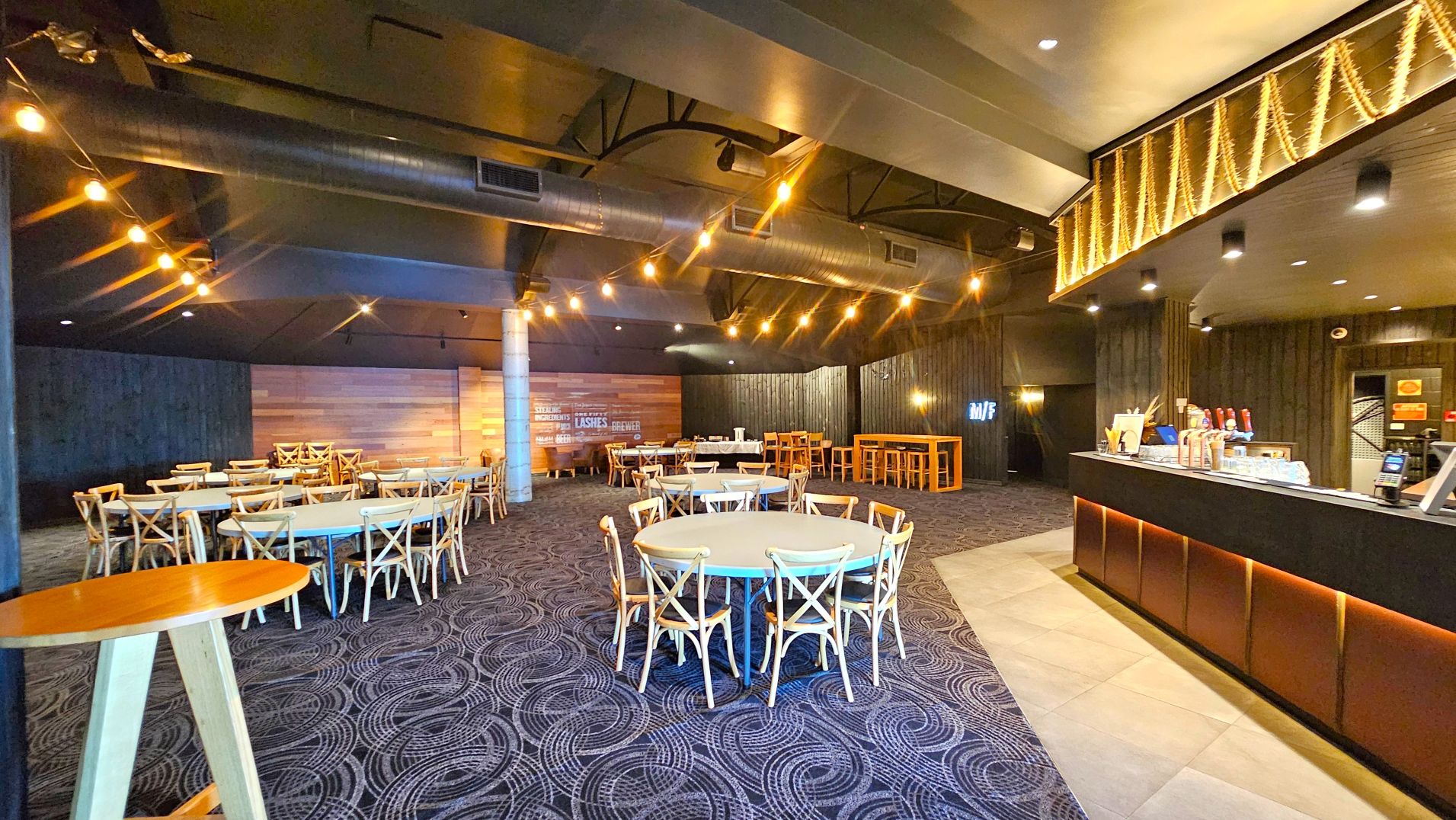 A spacious indoor venue with a modern design featuring round tables surrounded by wooden chairs on a patterned carpet. A long bar counter with a dark finish and illuminated base is on the right, with various taps and a payment terminal. The ceiling has exposed ductwork and string lights. Wooden panelling decorates the walls, and a small section of tiled flooring is visible near the bar.