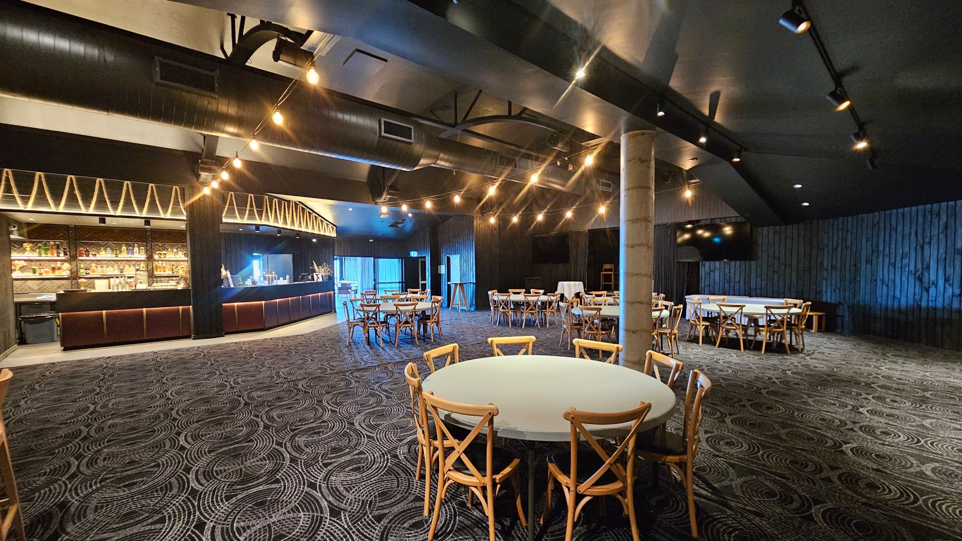 A spacious indoor venue with a modern bar area on the left, featuring shelves stocked with bottles and a sleek counter. The ceiling is adorned with exposed ductwork and string lights. Several round tables with wooden chairs are scattered across the patterned carpeted floor. A large pillar stands centrally, and the walls are dark with a textured finish. The space feels open and is designed for social gatherings.