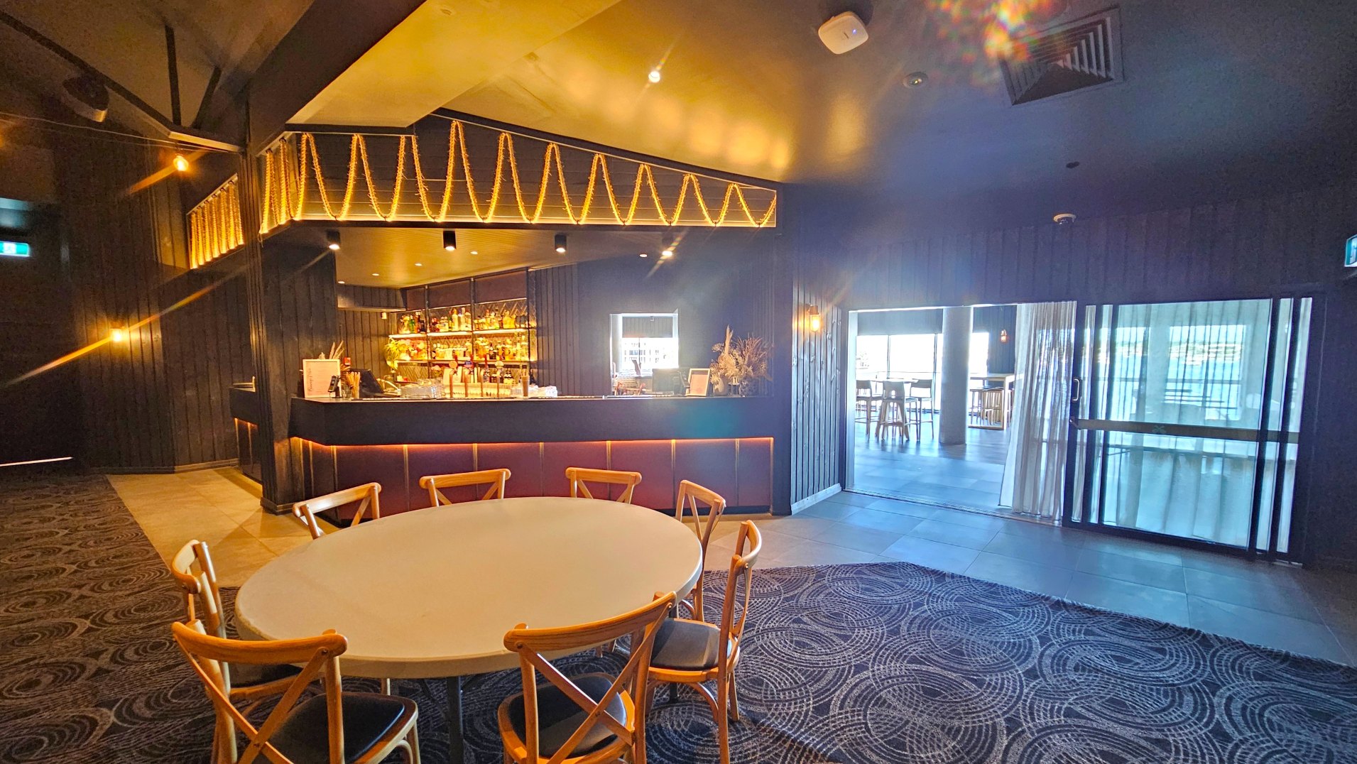 The image shows a modern bar area with a dark wood finish and warm lighting. A round table with wooden chairs is in the foreground on a patterned carpet. The bar counter is illuminated with string lights above, and shelves stocked with bottles are visible behind. To the right, a glass sliding door leads to a bright area with high tables and stools. The floor transitions from carpet to tiles near the door.