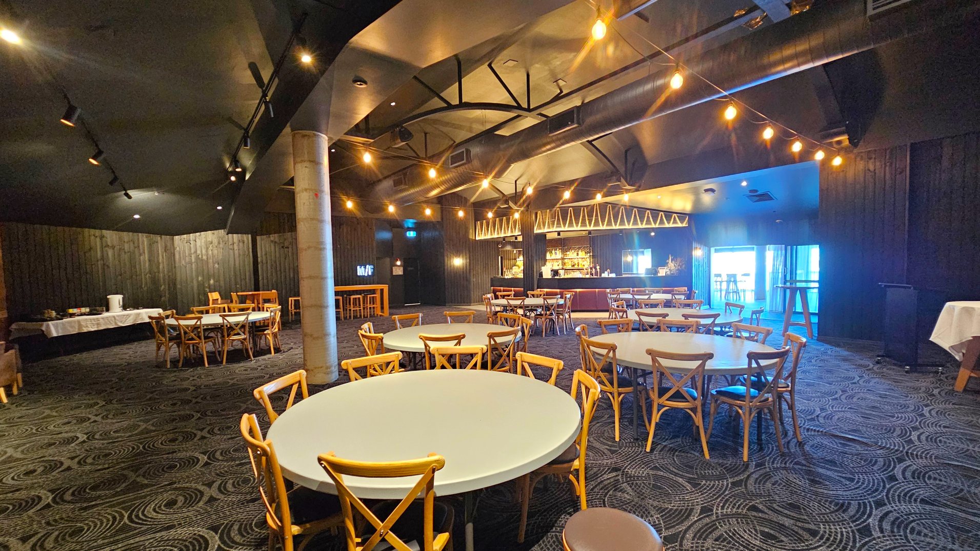 A spacious indoor venue with round tables and wooden chairs arranged on a patterned carpet. A prominent bar with a geometric design and illuminated shelves is visible in the background. The ceiling features exposed beams and string lights, adding a warm glow. A concrete pillar stands centrally, and a table with a white cloth is set against a dark wooden wall. A doorway with a blue light leads to another area.