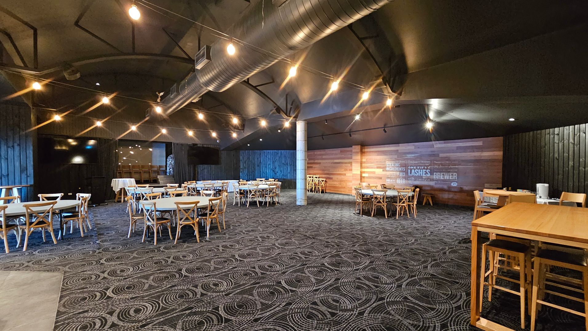A spacious indoor venue with a patterned carpet featuring circular designs. The room is furnished with wooden tables and chairs arranged in clusters. A prominent metal duct runs across the ceiling, which is adorned with string lights. The walls are a mix of dark wood panelling and lighter wood with signage. A concrete pillar stands centrally, and a high wooden table with stools is visible on the right.