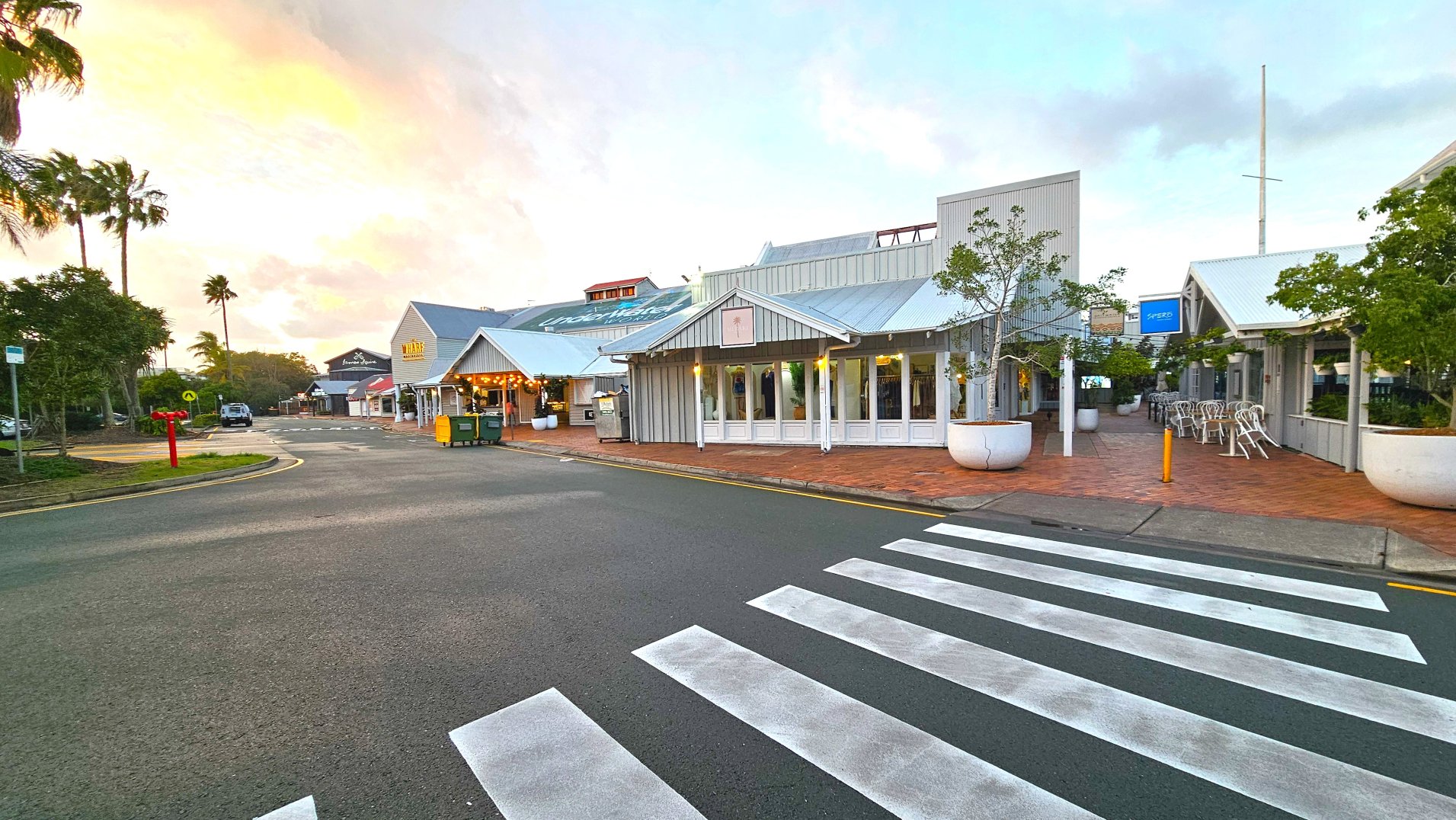 A street scene featuring a pedestrian zebra crossing leading to a complex of low-rise buildings with corrugated metal roofs and wooden facades. The buildings house various shops and eateries, with outdoor seating visible. Large potted trees line the brick-paved walkway. A few palm trees are in the background, and a yellow bin is near the entrance. The area feels open with a clear view of the surroundings.