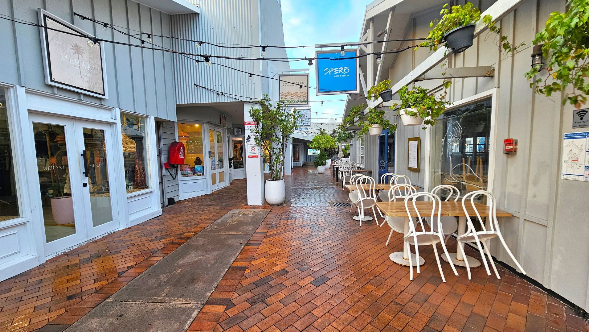 A narrow pedestrian alley with brick paving, flanked by shops and a café. On the left, a shop with large glass doors and a sign above. On the right, a café with white chairs and wooden tables under hanging plants. Overhead, string lights are suspended. A blue sign for 'Spero Greek Street' hangs above. The alley is enclosed by buildings with grey and white panelling, creating an intimate atmosphere.