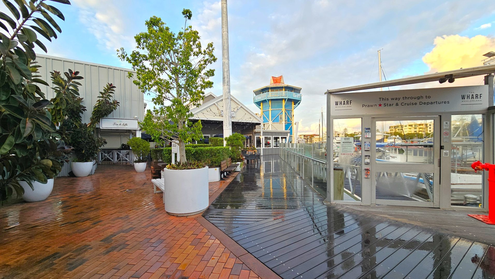 A waterfront area with a mix of brick and wooden decking surfaces. On the left, large white planters with lush green plants line the path leading to a building with a sign reading 'Fior di Latte'. A prominent blue and yellow tower stands in the background. To the right, a glass entrance with signage for 'Wharf' and 'Prawn Star & Cruise Departures' is visible, with boats docked beyond.