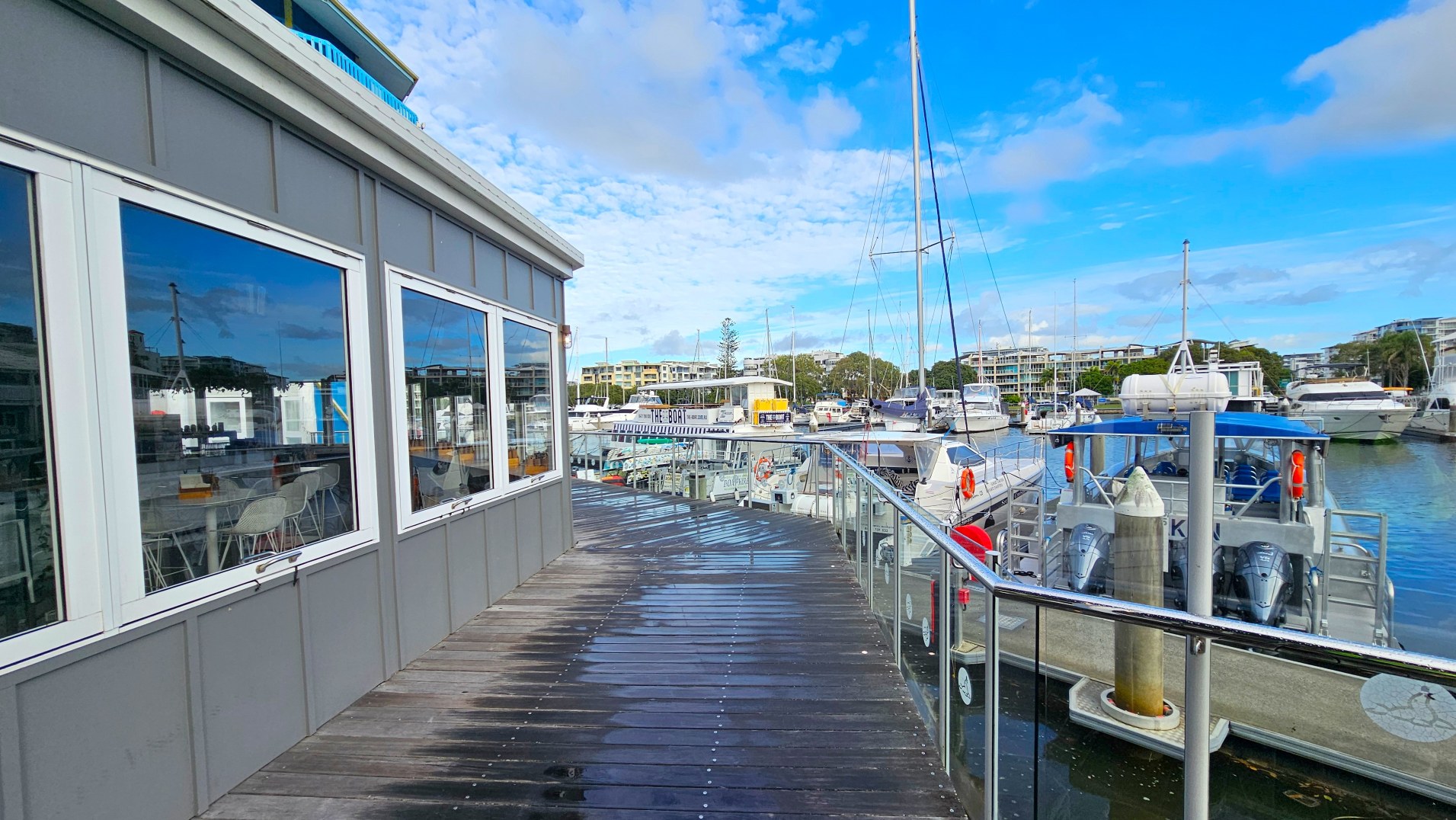 A waterfront scene featuring a wooden boardwalk with glass railings leading past a building with large reflective windows. The boardwalk is slightly wet, suggesting recent rain. To the right, several boats are moored, including a prominent boat with a blue canopy. In the background, modern buildings and trees are visible, creating a marina atmosphere. The area feels open with clear views across the water.