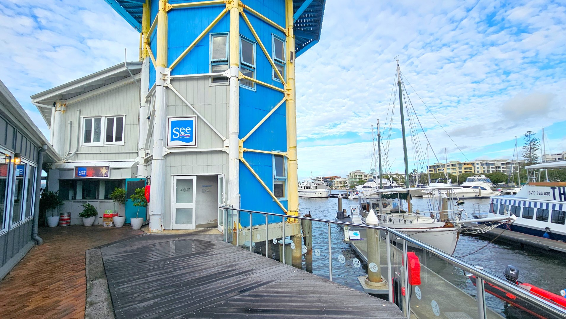 A waterfront scene featuring a two-storey building with a blue and grey facade, supported by yellow beams. The building has a sign reading 'See Restaurant'. A wooden boardwalk with a metal railing runs alongside, leading to a marina filled with various boats and yachts. Potted plants are placed near the entrance of the building. The area feels open with views of distant buildings across the water.