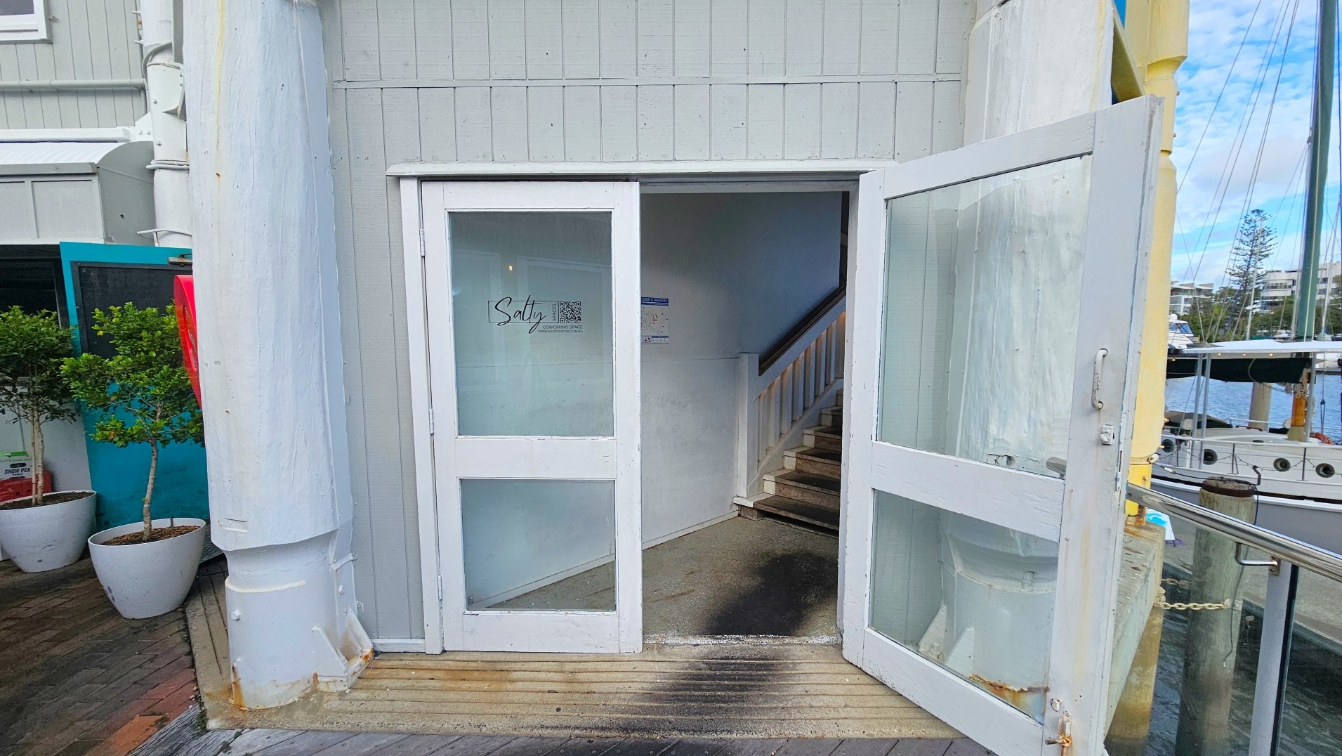 A white-framed glass door is open, leading into a narrow interior space with a staircase ascending to the right. The exterior wall is clad in light grey vertical panels. A sign on the door reads 'Salty'. The entrance is bordered by a wooden deck with a metal railing on the right, overlooking a marina with boats. Two potted plants are visible to the left, and a large white structural column stands adjacent to the door.