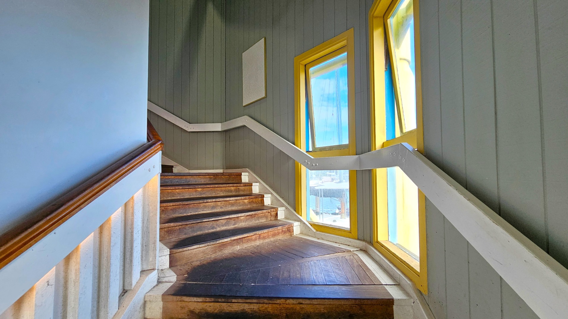 A wooden staircase with a dark brown finish ascends to the left, bordered by a white handrail. The walls are clad in vertical grey panelling, and three large windows with yellow frames allow natural light to flood in, casting shadows on the steps. The floor at the base of the stairs is a continuation of the wooden material, transitioning smoothly from the steps. A blank white rectangular sign is mounted on the wall above the stairs.