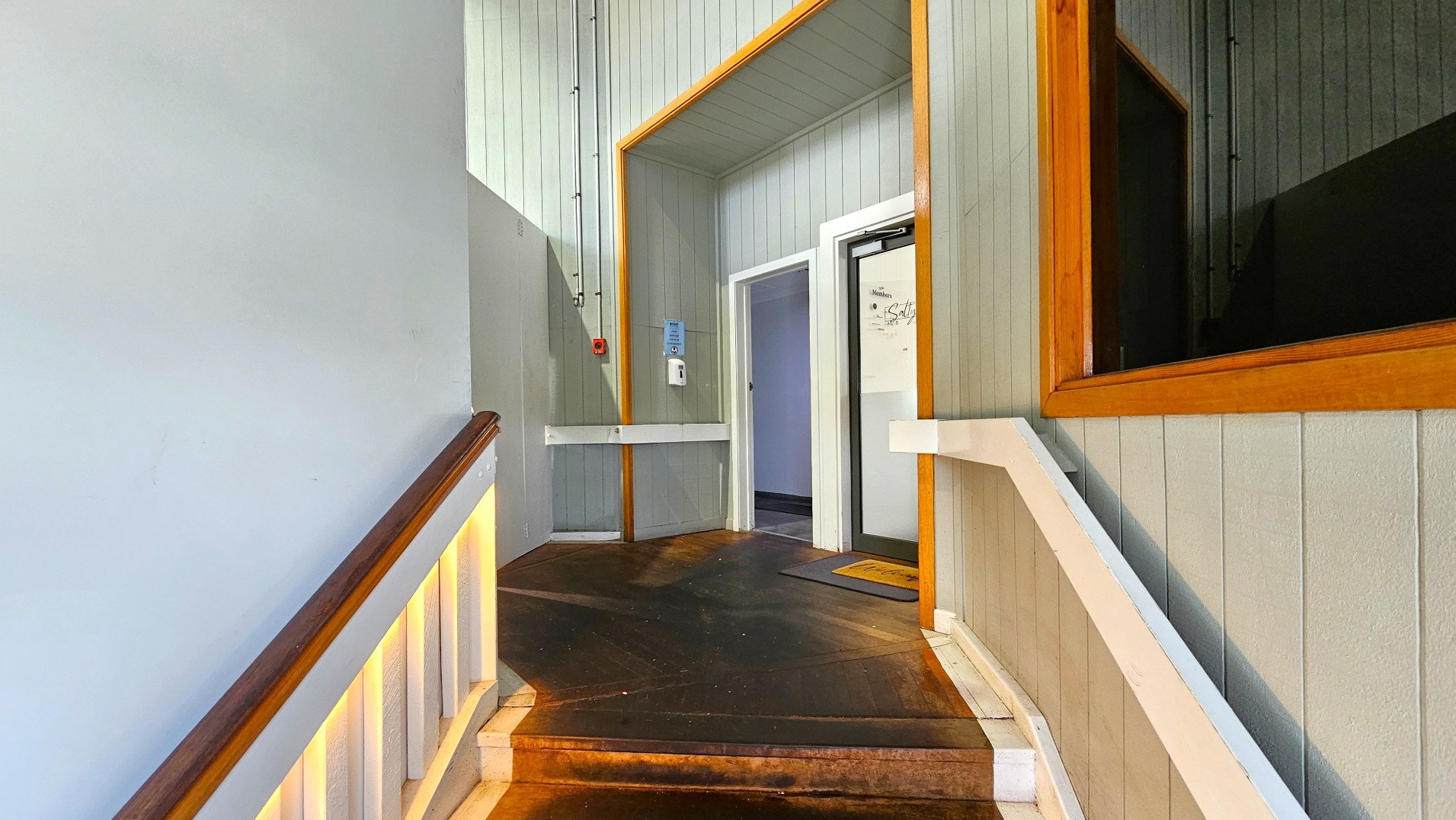 A narrow corridor with light grey panelled walls and a wooden handrail on the left. The floor is a dark wood, transitioning to a lighter wood at the entrance of a room. A white door with a glass panel is on the right, framed by a wooden border. Above the door, a small sign reads 'Salty'. A wall-mounted hand sanitiser and a red emergency button are visible on the left wall.