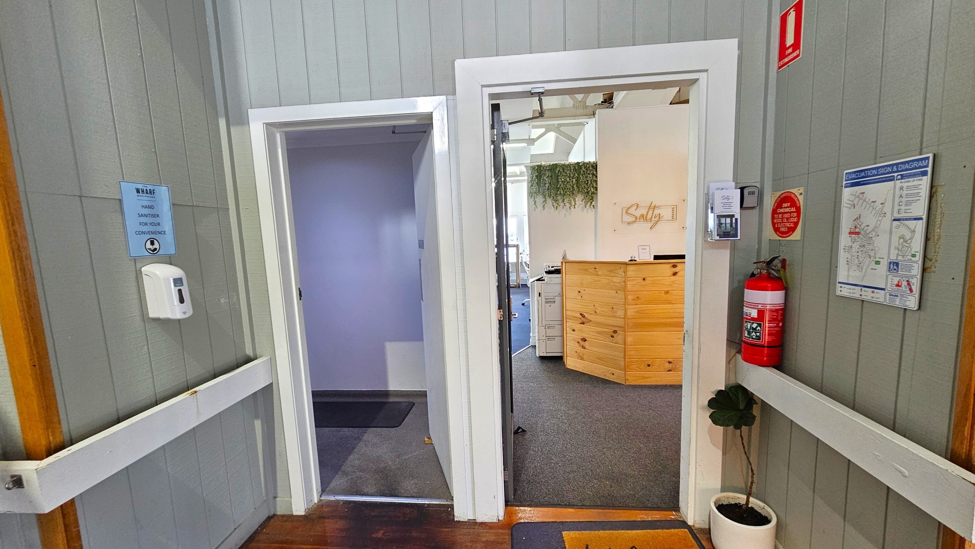 A hallway with grey panelled walls features two open doorways. The left doorway leads to a dimly lit room with a dark carpet. The right doorway opens into a brighter space with a wooden reception desk and a sign reading 'Salty'. A fire extinguisher and evacuation diagram are mounted on the wall. A hand sanitiser dispenser is affixed to the left wall. The floor transitions from wood to carpet at the doorways.