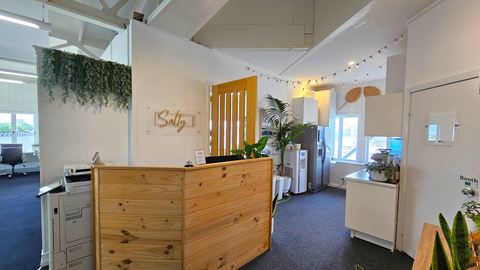 An office reception area with a wooden front desk adorned with potted plants. Behind the desk, a sign reads 'Salty'. To the right, a kitchenette with a fridge, water cooler, and coffee machine is visible. The space is decorated with string lights and hanging plants. A door marked 'Booth' is on the right. The area is carpeted, and large windows provide natural light. A printer is positioned to the left near an open workspace.