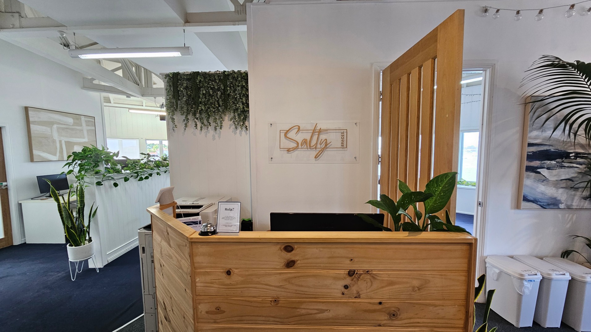 A reception area with a wooden desk featuring a small sign and a bell. Behind the desk, a wall displays a sign reading 'Salty' in cursive. A wooden door is partially open, leading to another room. Potted plants are placed around, and a hanging plant decorates the wall. The floor is carpeted in dark blue, and there are white walls with framed artwork. Overhead, fluorescent lights are visible.