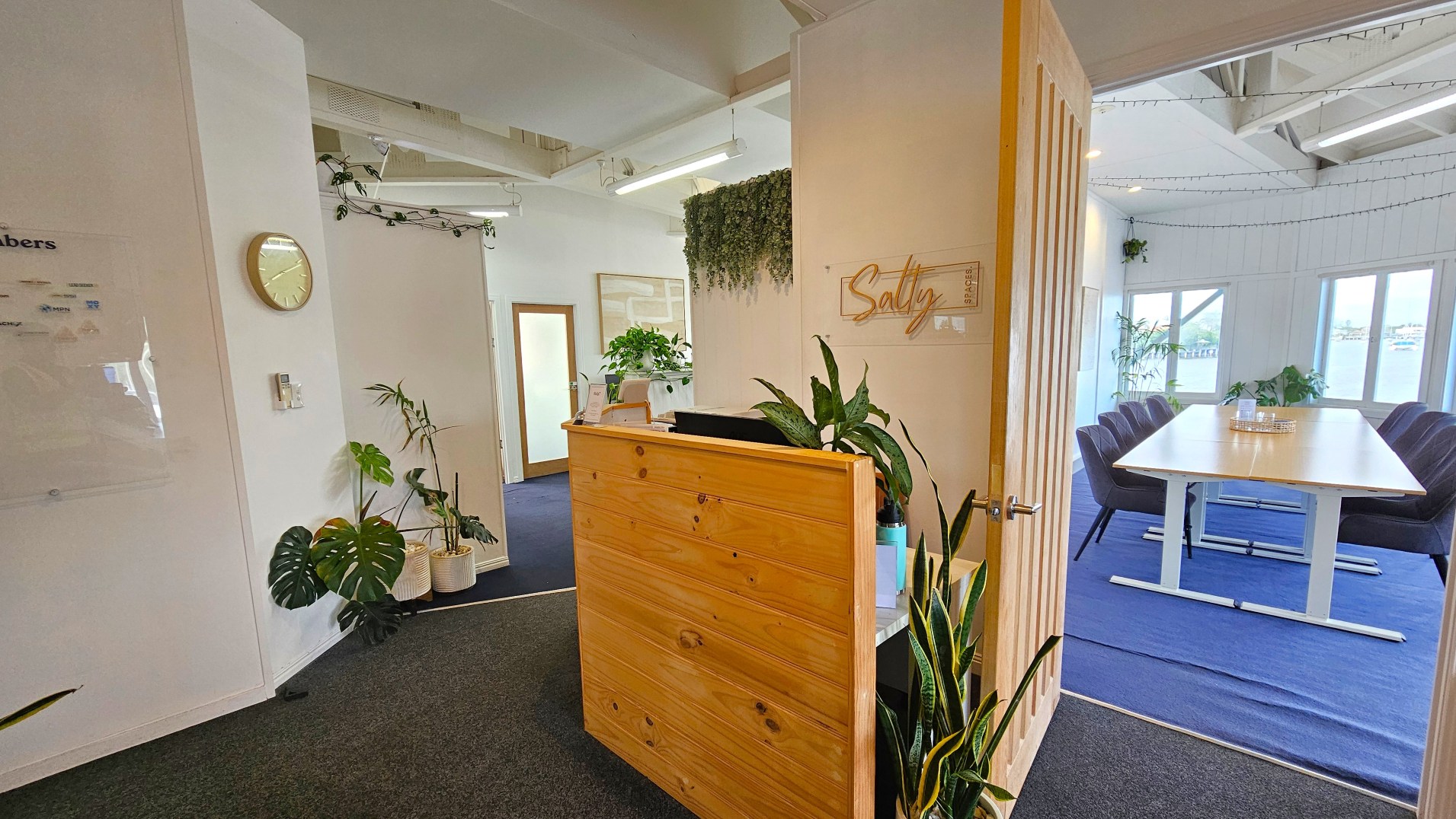 The image shows a modern office space with a wooden reception desk adorned with plants. A sign reading 'Salty' is mounted on the wall behind. To the right, an open door leads to a meeting room with a long table surrounded by grey chairs on a blue carpet. The room has large windows and is decorated with more plants. The walls are white, and a clock is mounted on the left wall. The ceiling features exposed beams and lighting fixtures.